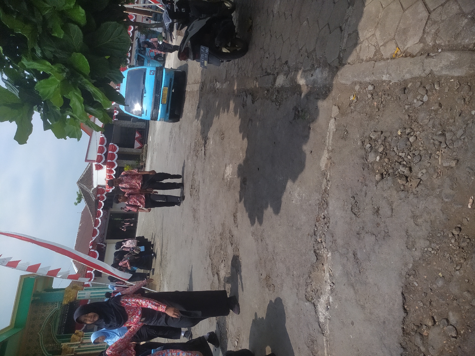 In the city of Candirejo, Indonesia, a group of students in school uniforms are gathered on a street with a rough asphalt surface. The students, mostly young females wearing hijabs and red and white patterned shirts with black skirts, are walking or standing. Two male students in similar uniforms are also visible walking together. In the background, a blue van is parked, and a motorcycle is seen on the right side of the image. Decorations, including red and white banners and flags, suggest a celebratory event. The environment appears to be an outdoor setting with buildings and trees.