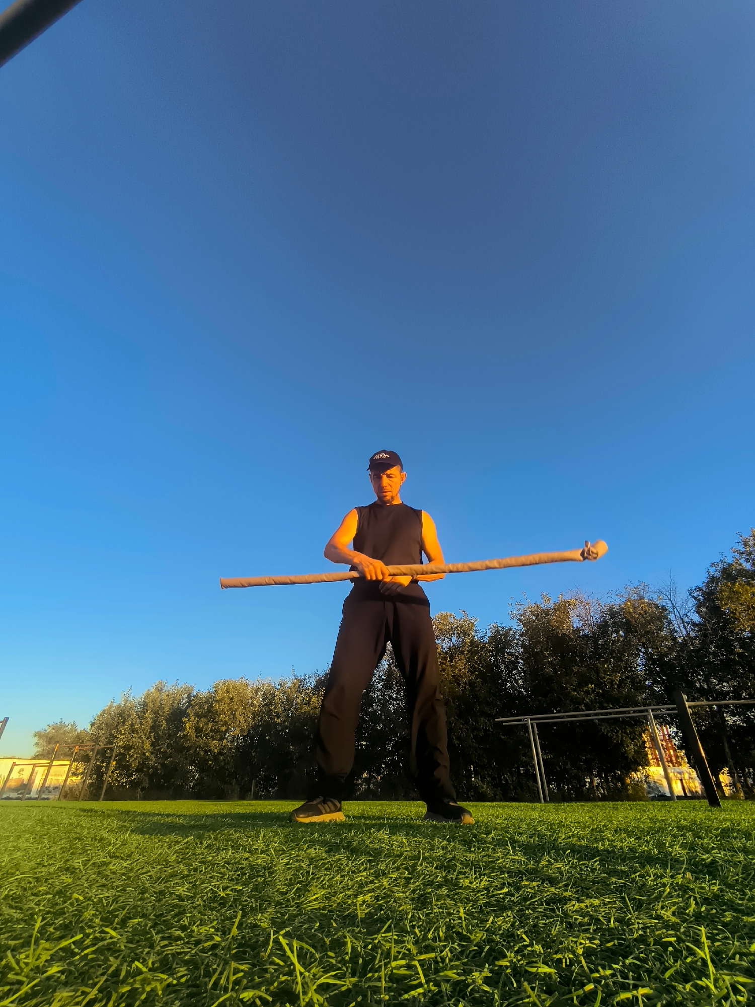 A young man wearing a black sleeveless shirt, black pants, a black baseball cap, and black athletic shoes stands on a grassy field under a clear blue sky. He is holding a long wooden staff horizontally in front of him with both hands. The staff appears to be wrapped in some sort of material at the ends. Behind him, there is a line of trees, and further back, there are metal structures that resemble sports equipment or fencing. The sun is low in the sky, casting long shadows on the grass and illuminating the man's arm from the side. The scene suggests an outdoor training or practice session. The location is identified as the city of Qarağandı, Kazakhstan.