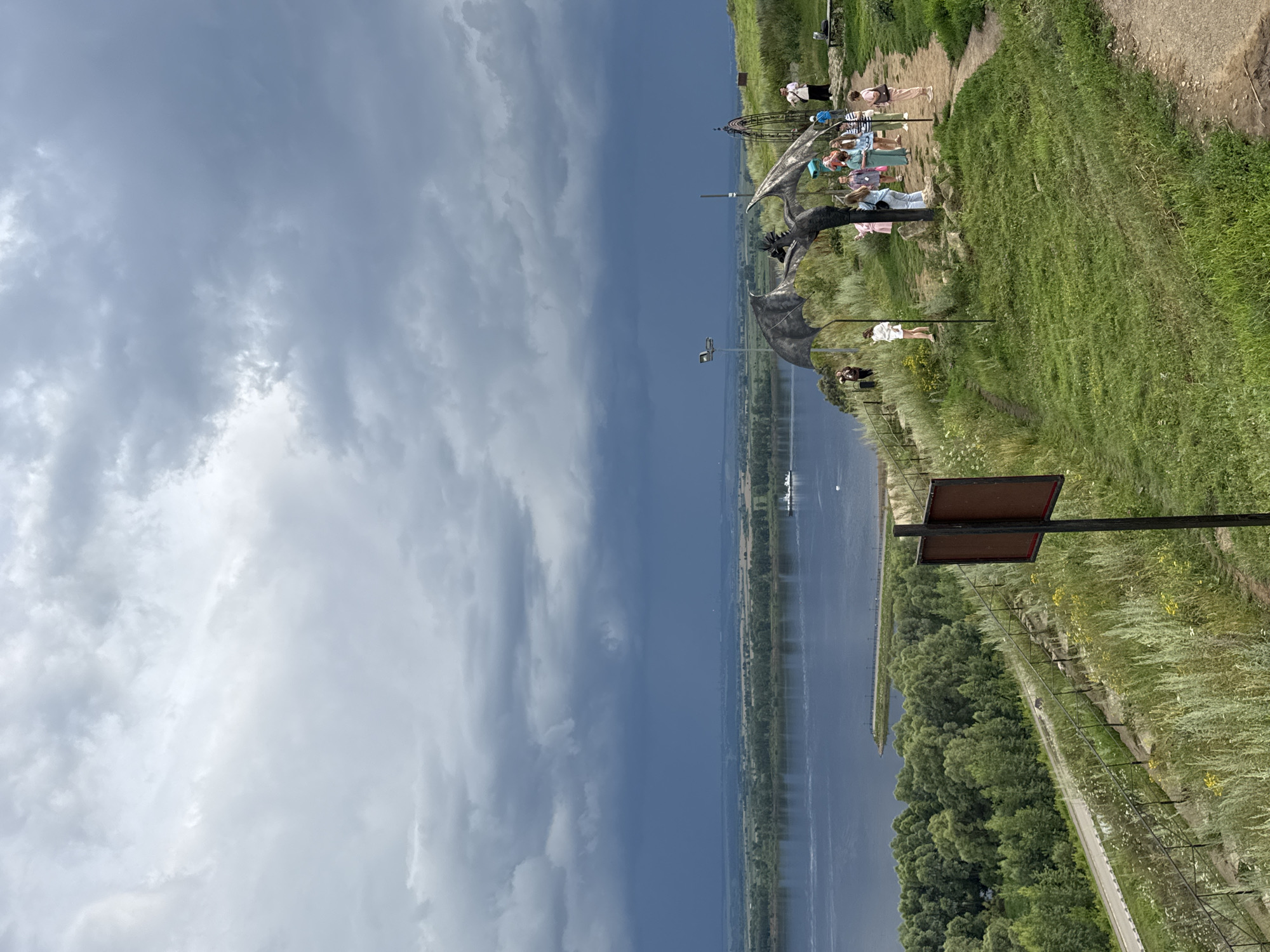 The image depicts a scene in Yelabuga, Russia, likely at the observation deck of the Shishkin Memorial Museum, overlooking the Kama River. The foreground features a grassy slope with a dirt path winding through it. Several people, including women and children, are gathered near a large, metallic sculpture of a winged creature, possibly a dragon. The sculpture is dark grey and intricately detailed, with outstretched wings. A tall, dark metal signpost with two rectangular brown panels stands to the left of the path.
Beyond the immediate foreground, the landscape unfolds to reveal the wide, calm expanse of the Kama River. A small white ferry or boat is visible on the water, moving from right to left. The riverbank is lined with dense green trees, creating a lush, natural border. In the distance, the land is relatively flat, with fields and more trees stretching towards the horizon.
The sky above is dramatic and overcast, with heavy, dark grey storm clouds dominating the upper portion of the image. Patches of lighter grey and white clouds are visible, suggesting the presence of a storm or heavy rainfall. The overall atmosphere is somewhat somber due to the cloudy sky, yet the presence of people and the river view suggest a popular tourist or recreational spot.