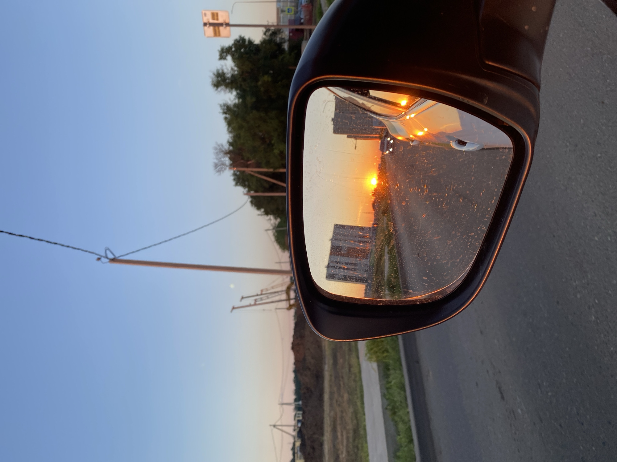 A side view mirror of a car reflects a sunset over a cityscape. The mirror shows the sun setting between two tall buildings, casting a warm glow on the scene. A car is visible driving on the road in front of the buildings, its headlights illuminating the sunset. The sky is clear and a pale blue, with a few wispy clouds. The reflection in the mirror is slightly distorted, giving the scene a dreamlike quality. The exterior of the car is visible in the lower right portion of the frame, its surface reflecting the orange glow of the sunset. The road in front of the car is asphalt, and there is a patch of green grass on the left side of the road.