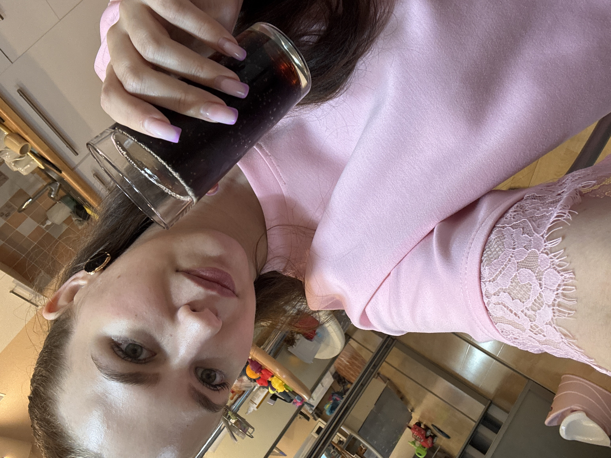 The image features a young adult female, positioned in a selfie-style shot, holding a clear glass filled with a dark, carbonated beverage. She looks directly at the camera with a neutral expression. Her hair is dark brown, and she wears a light pink long-sleeved top with a decorative lace cuff on the visible left sleeve. A gold-toned earring with a dark, irregular stone is present in her left ear. Her fingernails exhibit an ombre French tip style, transitioning from clear/pink to purple.
The background reveals an indoor domestic environment, likely a kitchen or dining area. Visible elements include light-colored wall tiles, a wooden shelf, and various household items in the background and reflected in a foreground surface. A pink pet bed or similar item and a child's toy are partially visible on the floor in the lower left quadrant. No specific geographic markers for Chaikovsky, Russia, are discernible within the image.