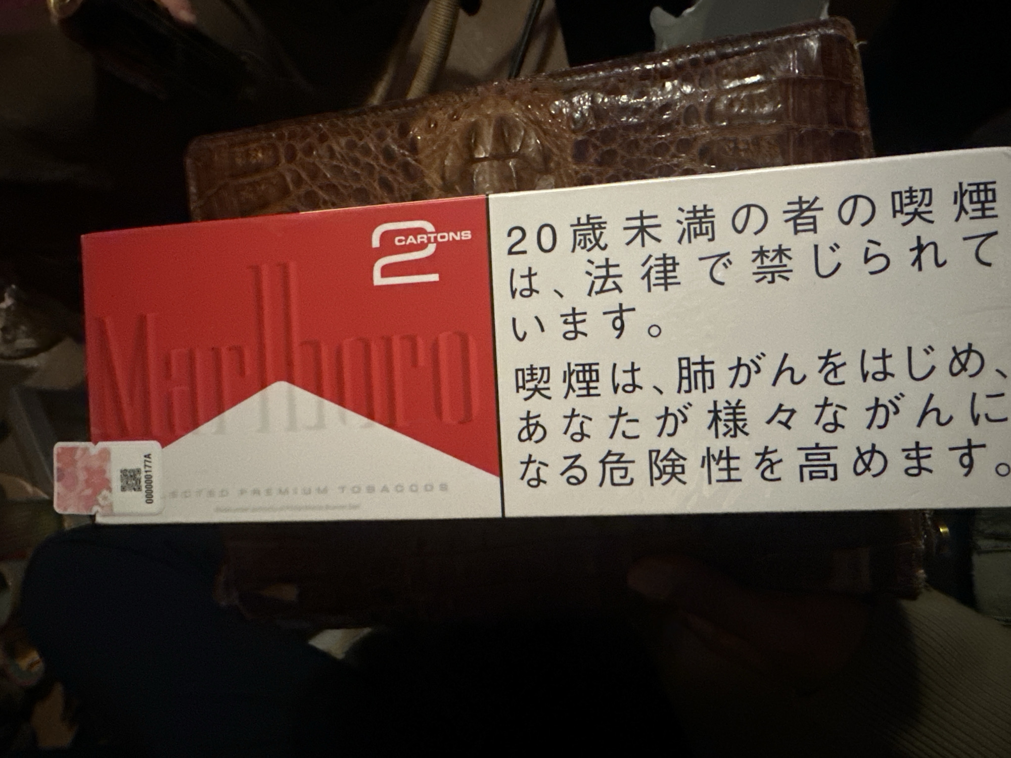 A pack of Marlboro cigarettes labeled "2 CARTONS" is resting on top of a brown leather wallet. The side of the pack displays a health warning in Japanese, indicating that smoking is prohibited for individuals under 20 years of age and increases the risk of various cancers. A serial number "000000177A" is visible on the side of the pack. The setting appears to be indoors, with a person wearing a blue shirt partially visible in the lower portion of the image. The exact location in Montreuil, France, is not identifiable from the provided visual information.