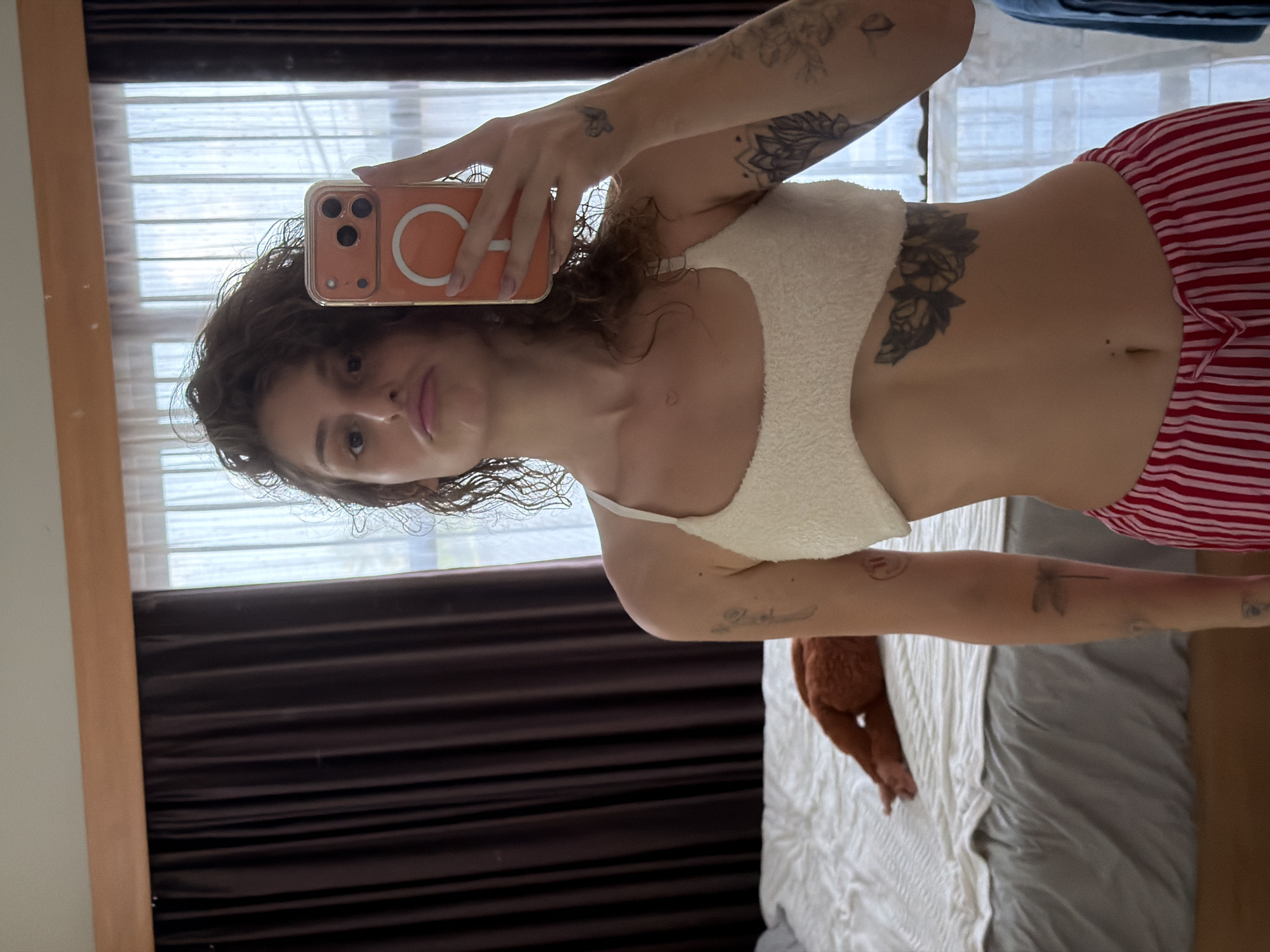 In a bedroom in North Kuta, Indonesia, a person is taking a mirror selfie. The individual is positioned in the foreground, holding an orange phone in their right hand. They are wearing a cream-colored bralette and red and white striped pants. Tattoos are visible on their arms and torso. The background features a bed with a brown stuffed animal on it, a dark brown curtain on the left, and sheer white curtains on the right. The room appears to be lit by natural light coming from the windows.