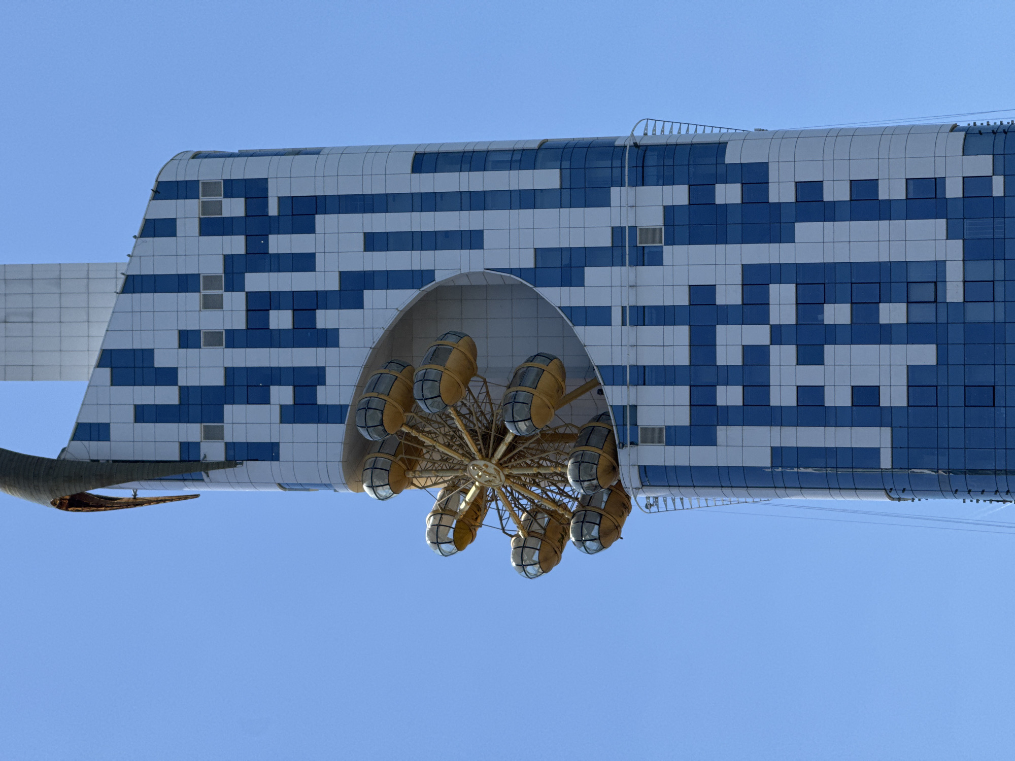 The image shows a low-angle view of the Alphabet Tower in Batumi, Georgia, set against a clear blue sky. The tower's unique architectural design features a latticework of blue and white square tiles. Attached to the tower is a Ferris wheel with golden carriages, suspended under an arched structure. Part of the tower's support structure, possibly another level or platform, is visible to the left. The angle emphasizes the height and architectural complexity of the tower.