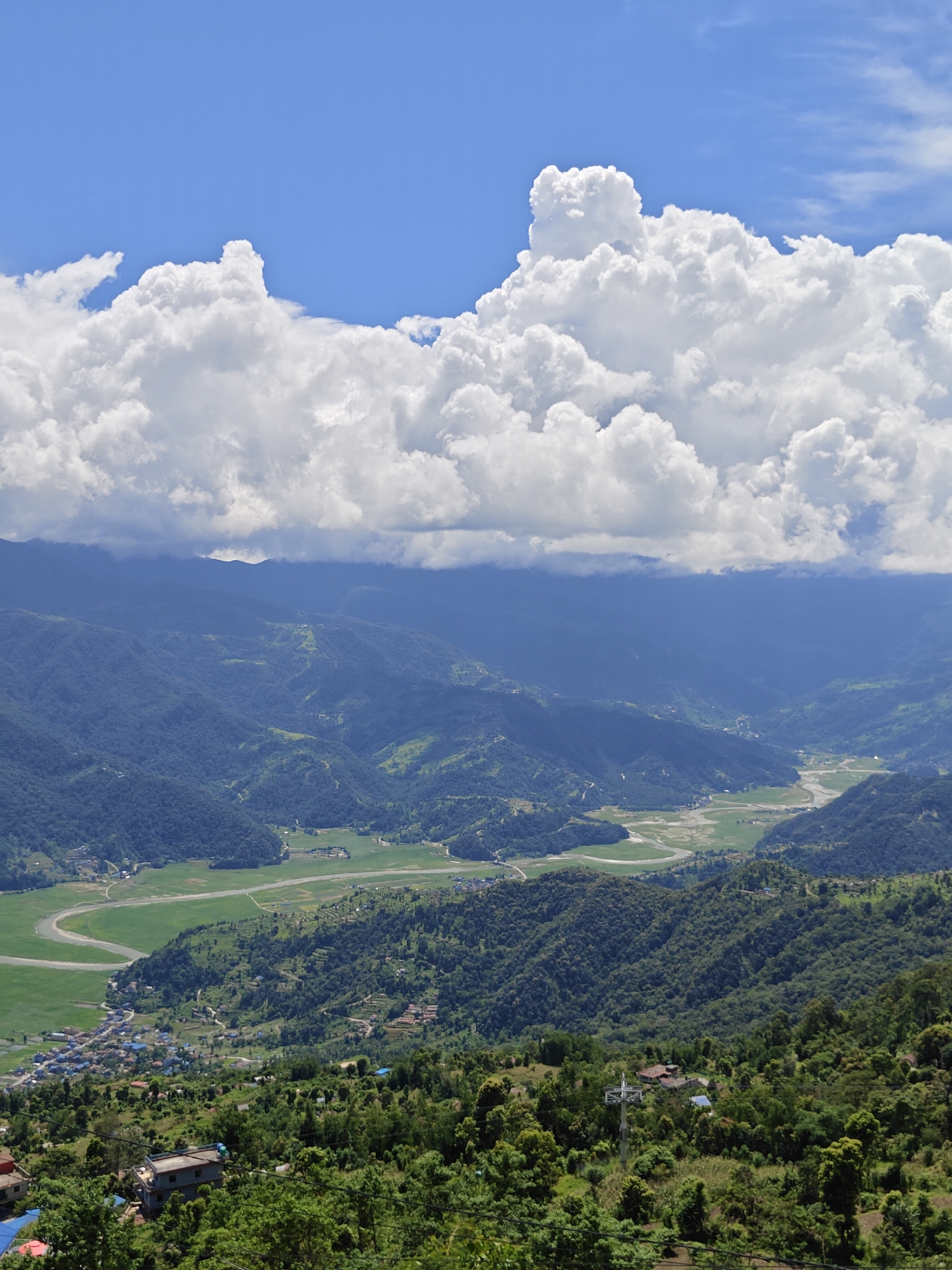 This is an aerial, full-frame, vertical shot of a lush, mountainous landscape under a partly cloudy blue sky, likely taken in or around Pokhara, Nepal. The foreground is dominated by dense, green foliage and scattered buildings, suggesting a hillside settlement. As the view recedes, rolling hills covered in dark green trees form a verdant carpet. Further in the distance, a wide, fertile valley stretches out, bisected by a meandering river or waterway. Agricultural fields, some appearing flooded or marshy, are visible in the valley floor. The mountains in the background are hazy and cloaked in a bluish-green hue, indicating their distance. The sky above is a vibrant blue, punctuated by large, white, puffy cumulus clouds, some of which cast shadows on the mountains. The scene conveys a sense of natural grandeur and rural tranquility.