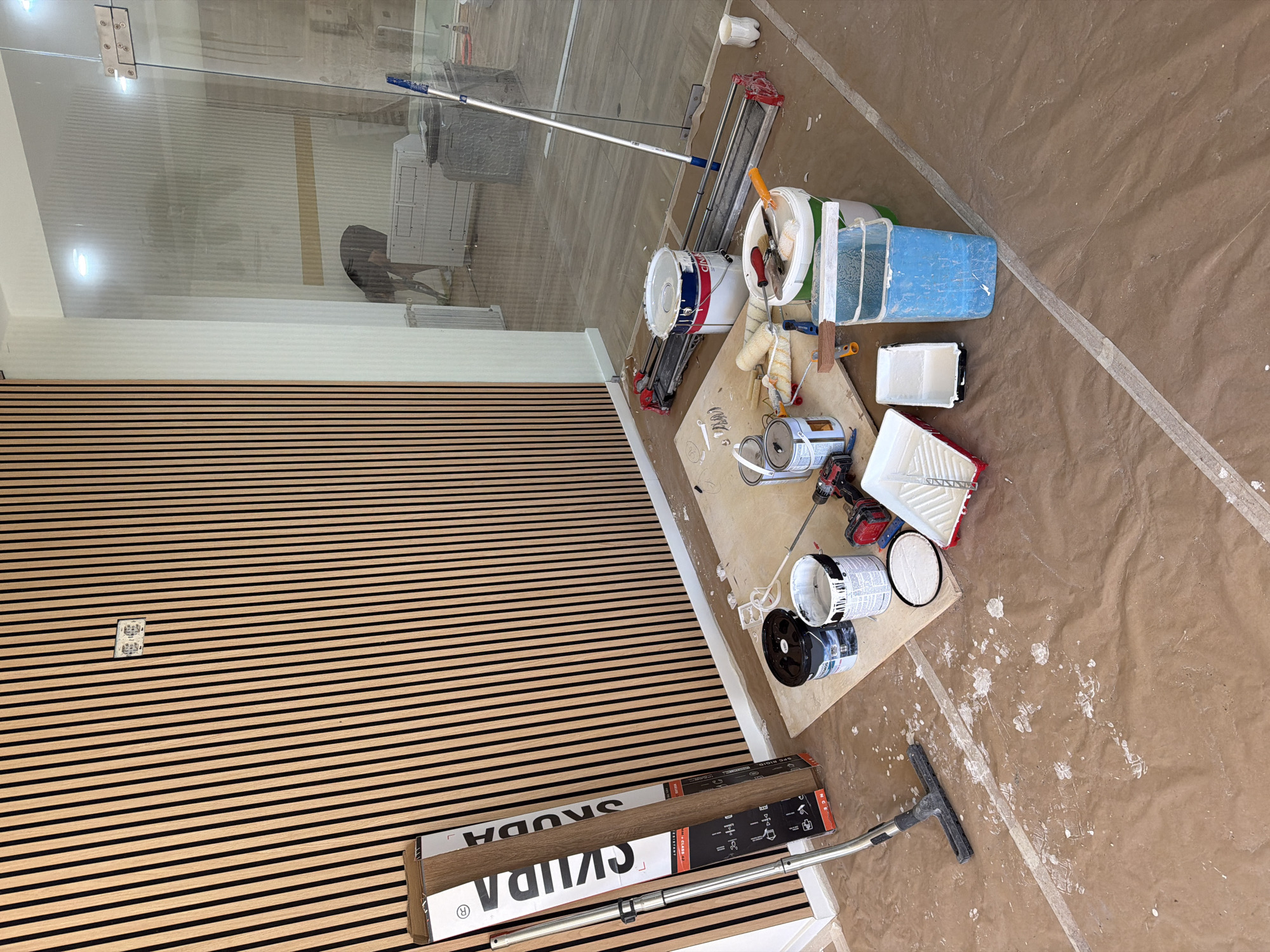 This image depicts a room undergoing renovation or construction, specifically located in Gondomar, Portugal. The scene is characterized by unfinished work and tools commonly associated with painting and tiling.
The walls are partially covered with vertical wooden slats, suggesting a modern design element. On the left side, a large cardboard box with the brand name "SKUDA" is propped against the slatted wall. Next to it, a silver pole with a brush attachment, likely a vacuum cleaner extension, lies on the floor.
The floor is covered with brown protective sheeting, splattered with white paint in various locations, indicating active painting work. Several paint cans are present, including a large white bucket with blue and red accents, another white can with a blue lid, and a smaller black can. Paint rollers, paint trays, and brushes are scattered around, some on a wooden board. A tile cutter is also visible on the floor.
In the background, through a glass partition, another part of the building is visible. This area appears to be a hallway or a larger open space with flooring and some furniture. There are also signs of ongoing work in that area, including what appears to be a person in the distance and a large grey bin. A long-handled mop or squeegee leans against the glass partition.
The overall impression is one of a work in progress, with the necessary tools and materials laid out for painting and possibly tiling. The lighting suggests it is daytime, and the environment is indoors.