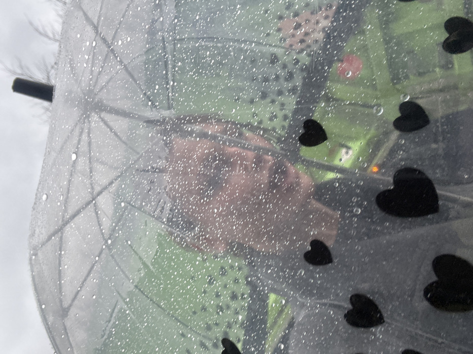 The image depicts an upward-angled view through a clear, rain-covered umbrella adorned with multiple black heart-shaped decals. Numerous water droplets are visible across the umbrella's surface, distorting the view. Behind the umbrella, the upper body and face of an individual with light-toned skin and dark clothing are partially visible, obscured by the rain and the umbrella's material. In the background, beyond the person, a prominent lime-green vehicle, consistent with a London bus livery, is discernible, with a faint orange light or reflection visible on its interior. The sky above the umbrella appears overcast, indicating rainy weather in an urban environment. The scene is located in the city of London, United Kingdom.