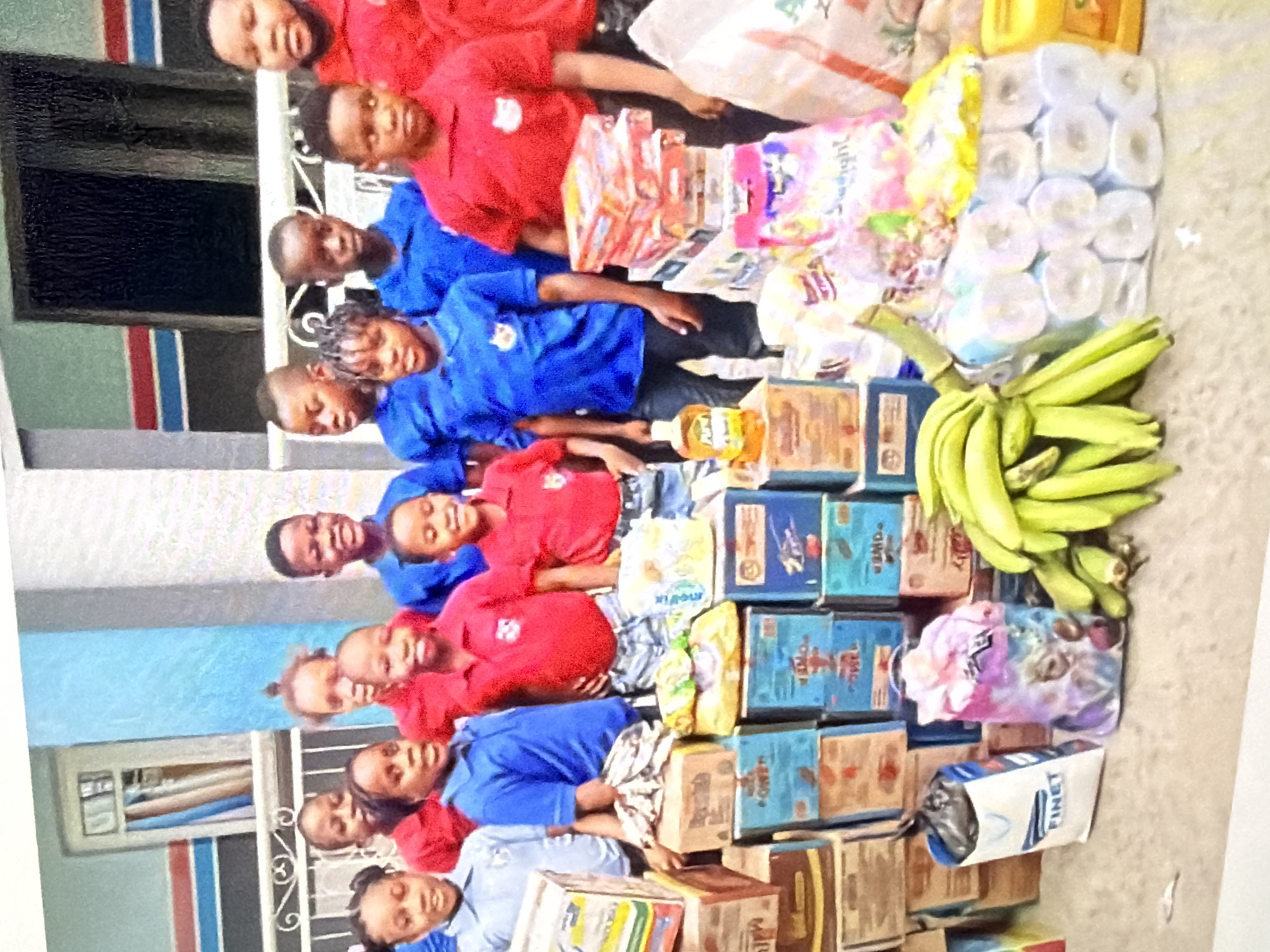 In an outdoor setting with a building facade in the background, a group of approximately ten Black children, all appearing to be elementary school age, stand behind a large assortment of stacked food items and supplies. The children are wearing blue or red collared shirts, possibly school uniforms, and some have smiles on their faces. In the foreground, numerous cardboard boxes are stacked, containing what appear to be food products. Among the boxes are also bags of what seem to be snacks and toilet paper rolls. A bunch of green bananas is also visible in the foreground. The overall scene suggests a distribution of goods to children.