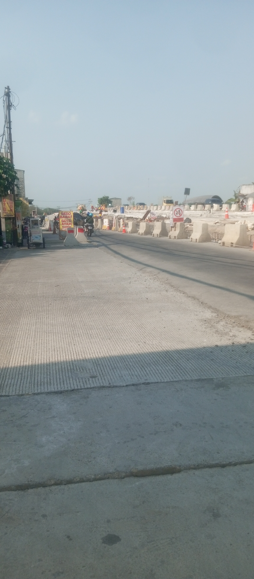 The image depicts a street scene in Kediri City, Indonesia, featuring ongoing road construction. The foreground shows a concrete road surface with distinct grooved lines, suggesting a newly laid or resurfaced section. In the middle ground, a motorbike is being ridden down the road, passing by temporary concrete barriers and construction materials that line the sides. Several traffic cones and construction signs are visible, indicating a designated work zone. Further in the background, more construction activity is apparent, with trucks and workers engaged in building a road or bridge. The sky is clear and bright, indicating daylight. The overall impression is one of active infrastructure development in an urban or semi-urban setting.