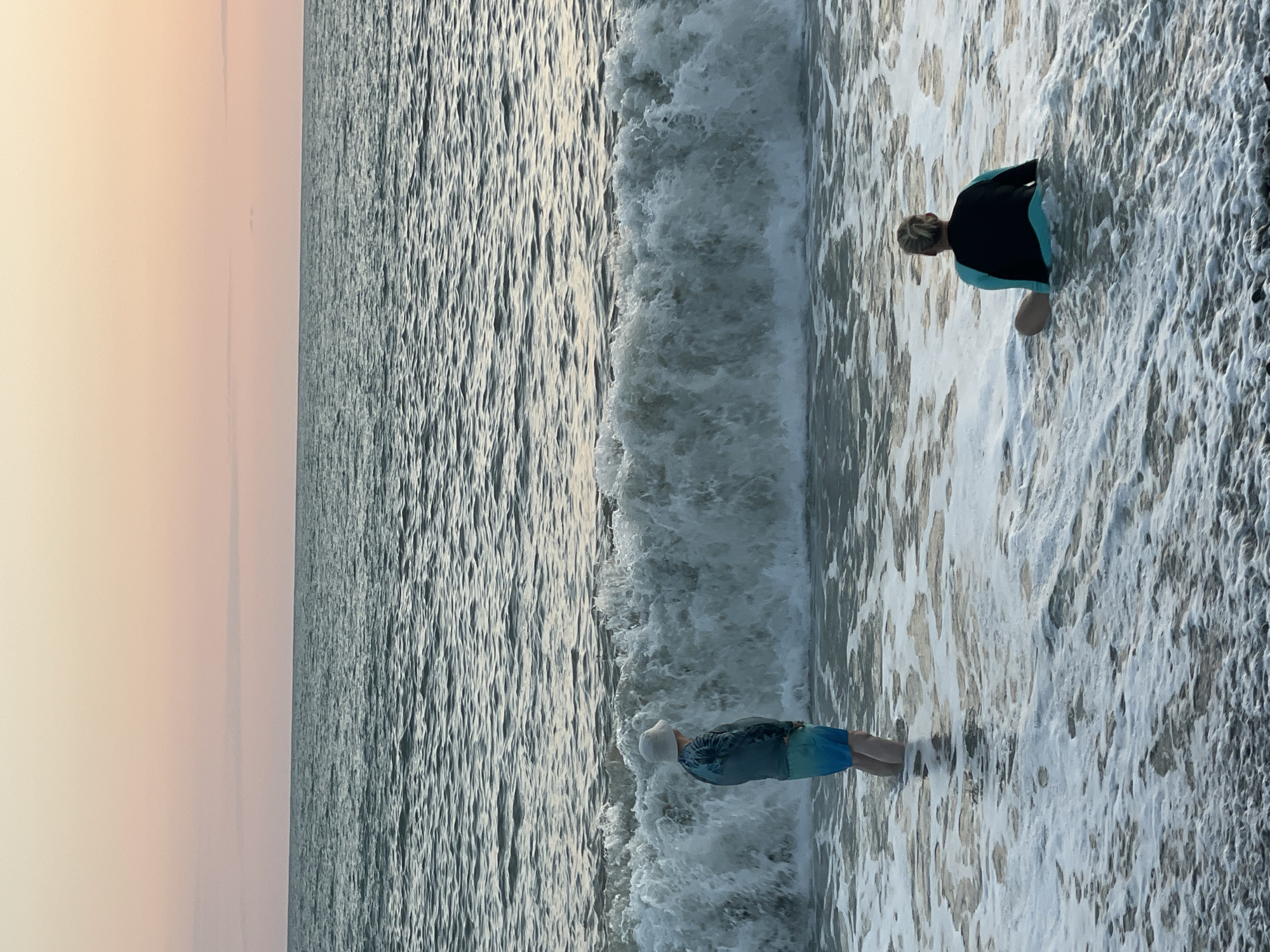 Two people are standing in the ocean on the coast of Sochi, Russia, at sunset. The person on the left is wearing a hat, a long-sleeved blue and green shirt, and blue and green swim trunks. They are standing in shallow water, facing the ocean. The person on the right is sitting in the water, wearing a black and light blue rash guard. They are facing away from the ocean. A large wave is breaking behind the person on the left. The sky is a soft pink and orange, indicating the sun is setting. The water is a dark blue-grey, with white foam from the waves. The beach in the foreground is made of dark pebbles.
