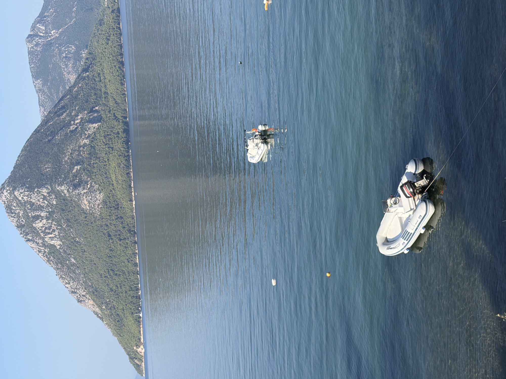 The image depicts a body of water with a mountainous coastline in the background. The water is a deep blue and has ripples on its surface. There are several inflatable boats in the water. One larger inflatable boat with two outboard motors is anchored near the shore in the foreground. Further out in the water, there are two smaller inflatable boats. In the far distance, a range of mountains covered in green vegetation rises from the coastline, with rocky peaks visible at the summit. The sky above is clear and blue. The scene appears to be a tranquil coastal area.