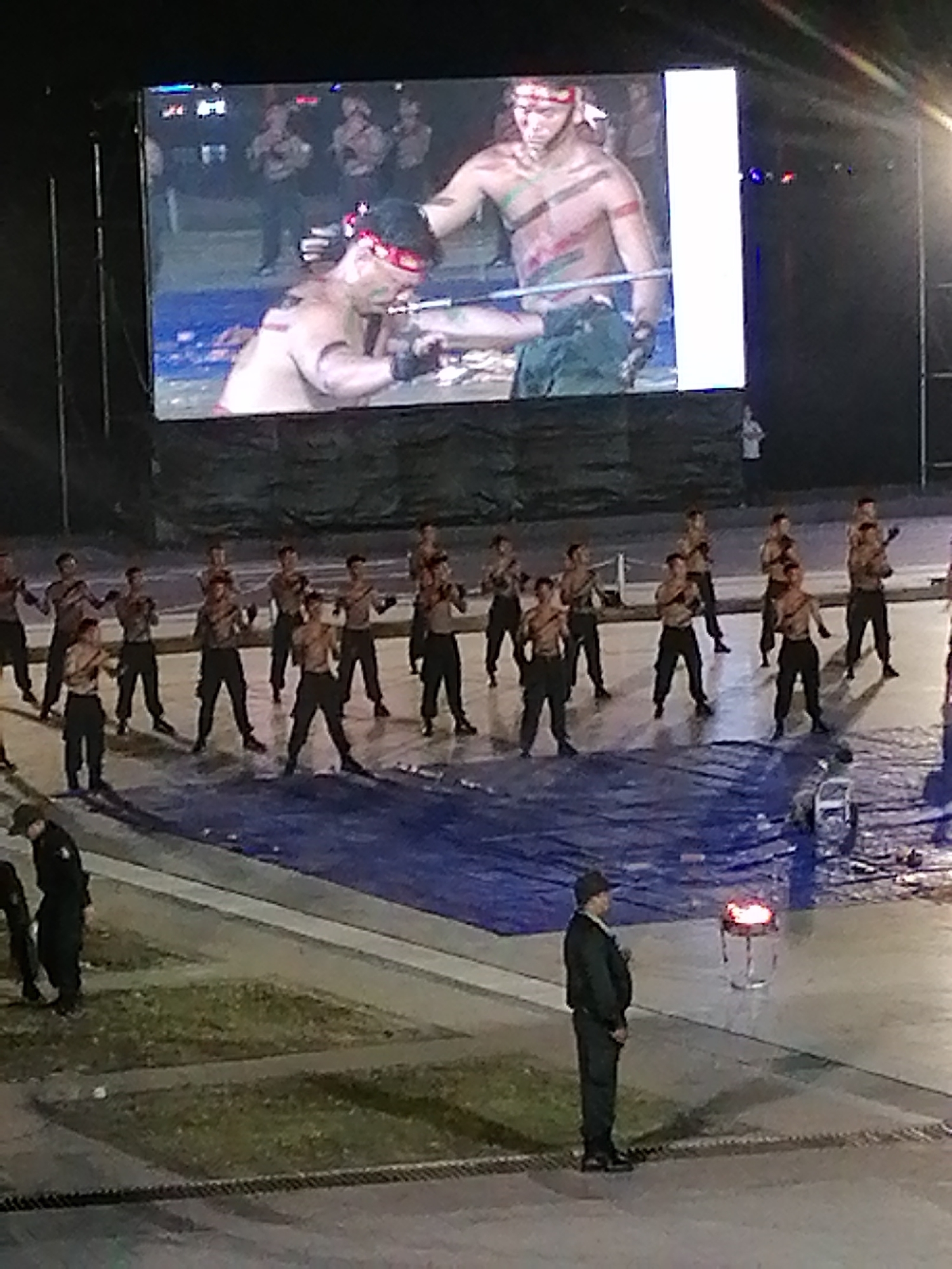 The media file shows an outdoor event at night, likely in Da Lat, Vietnam, given the location context. In the foreground, two individuals are visible. One is wearing a dark uniform with a hat, standing at attention. Another person is partially visible to the left, also in dark attire.
The main focus of the image is a large screen displaying a performance. On the screen, two men are depicted in what appears to be a martial arts or combat demonstration. Both men are shirtless and appear to have body paint or markings on their chests and arms, suggesting a cultural or ritualistic performance. One man is striking a defensive pose, while the other is extending his arm, possibly in an offensive move. A group of other men, also shirtless and wearing dark pants, are standing in formation on the ground below the screen, mirroring the poses seen on the screen. They are arranged in rows and appear to be participating in the same performance.
The scene is illuminated by stage lighting, casting a glow on the performers and the large screen. The ground is covered in a blue tarp or fabric, with some debris visible. In the mid-ground, a few additional figures are seen, including one in a light-colored shirt and dark pants, and another in a dark uniform. A small, illuminated object resembling a beacon or lamp is present in the mid-ground to the right.
The overall atmosphere appears to be one of a lively evening event, possibly a festival or cultural showcase. The lighting and the dynamic poses of the performers suggest an energetic performance. No discernible text is visible in the image.