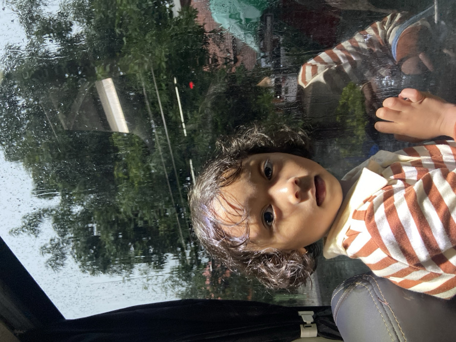 A young child with dark, curly hair is positioned in the foreground, seated within a vehicle and looking directly forward. The child wears a white-collared shirt with horizontal orange and white stripes. Their right hand is pressed against a side window heavily streaked with raindrops. To the left, a dark vehicle seat and a black curtain with a white clip are visible. The exterior view through the rain-covered window features dense green foliage from trees, partially obscuring a background containing indistinct urban elements, including a linear white structure and hints of a reddish building. The child's reflection, wearing the striped shirt, is visible on the wet glass surface. The scene is located in Pandaan, Indonesia.