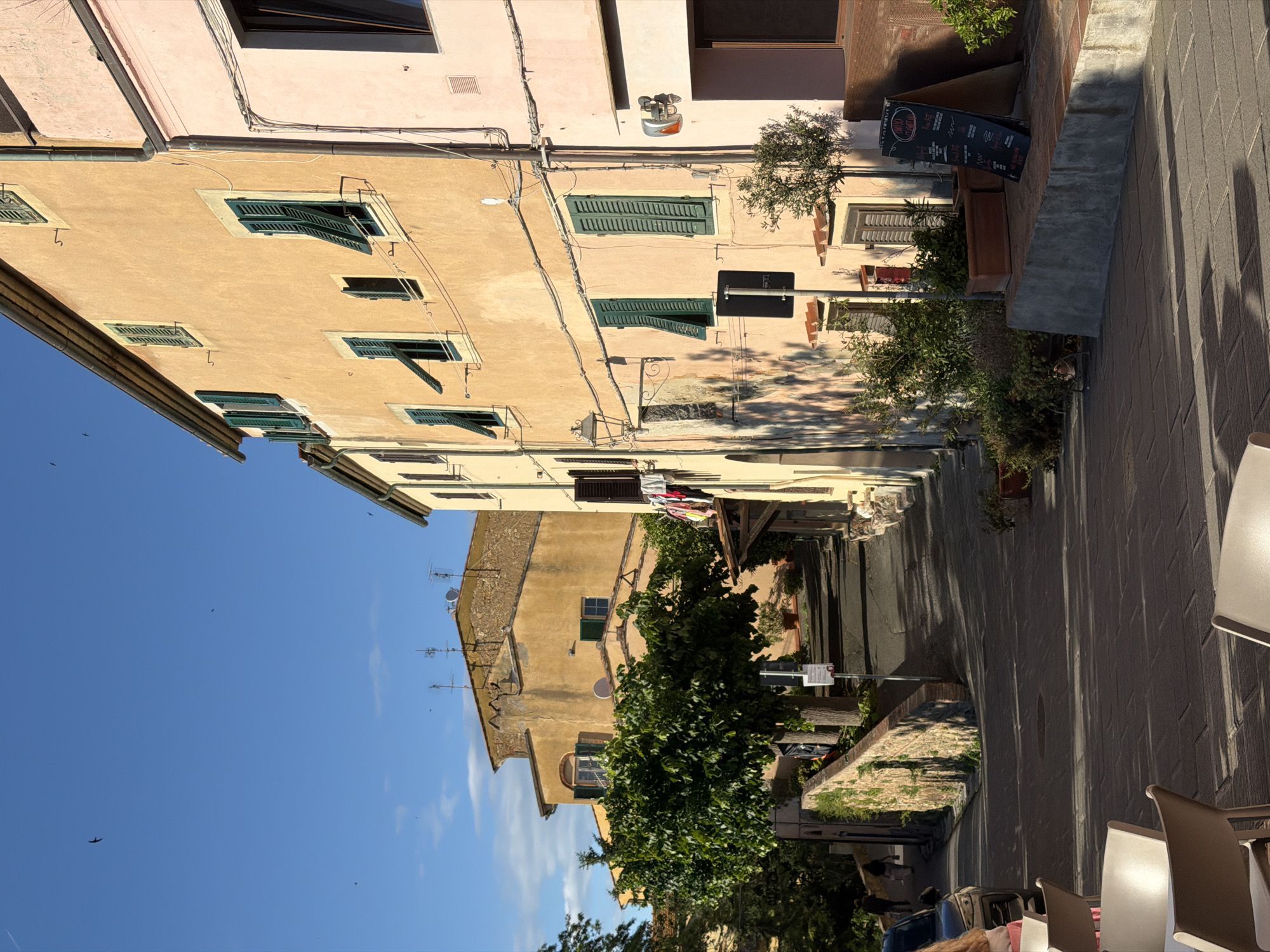 The image shows a street scene in Castagneto Carducci, Italy. Buildings line both sides of the narrow street. The buildings are constructed of stone and plaster, with traditional architectural details such as green shutters on the windows. Some laundry is hung out to dry between buildings across the street. The street is paved with stone and brick and is mostly in shadow. Tables and chairs for an outdoor cafe are visible in the foreground. A menu board stands outside a restaurant. There is vegetation along the street, including trees and potted plants. The sky is blue and mostly clear.