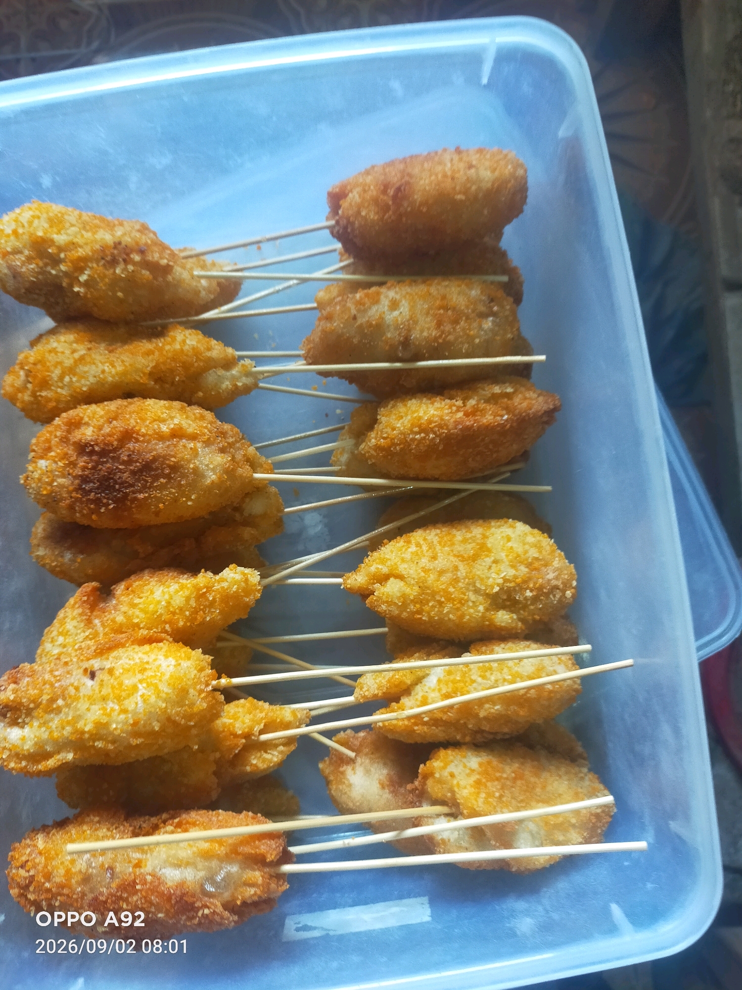 A close-up, overhead shot shows a clear plastic container filled with several fried, breaded food items on skewers. The food items appear to be small, cylindrical, and golden brown, suggesting they are deep-fried. They are arranged in vertical stacks within the container, with wooden skewers protruding from each. The background is blurred but hints at a tiled surface and a dark, possibly fabric, material. The image includes a timestamp at the bottom left: "OPPO A92 2026/09/02 08:01". The location is identified as Binangun, Indonesia. There are no people or visible actions in the frame.