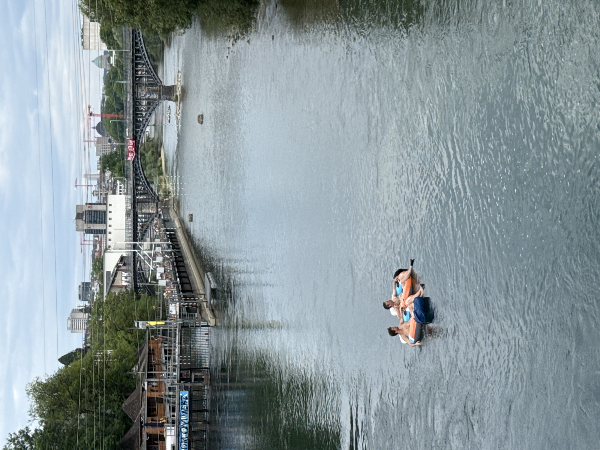This is a high-angle, full shot of a river flowing through Zurich, Switzerland, with a city skyline and bridge in the background. In the foreground, three people are floating in inner tubes. Two men are in one tube, one of whom appears to be sitting up and looking to the left, while the other is lying back and looking to the right. A third person, partially obscured, is in another tube to their right, also lying down. A blue bag is in the tube with the two men. The river is a deep blue-grey, with ripples and small waves. The background features a wooden structure on the left bank of the river, a metal bridge spanning the river, and various buildings and construction cranes in the distance. The sky is overcast.