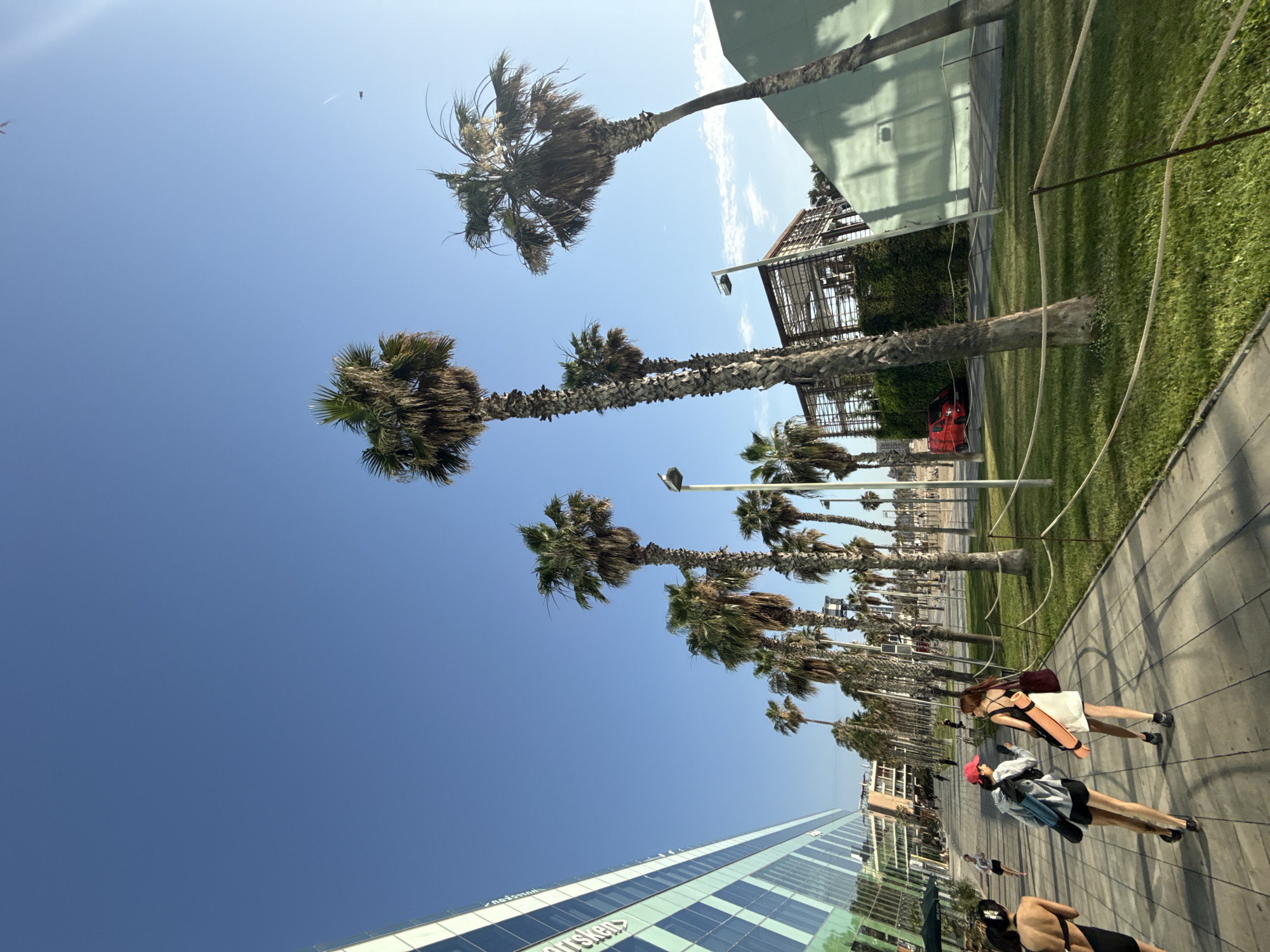 The image depicts a sunny day in Barcelona, Spain, likely near the beach or a waterfront area, given the presence of numerous palm trees and the pedestrian walkway. In the foreground, three individuals are walking away from the camera on a paved path lined with grass and rope barriers. The person on the far left, seen from the back, is wearing a black bikini. The person in the middle, wearing a red baseball cap and a grey hoodie over black shorts, is looking at their phone while carrying a blue rolled-up mat over their shoulder. To their right, a person in a white dress with a maroon bag slung over their shoulder is also walking.
To the left of the walkway is a modern, multi-story building with a glass facade, reflecting the blue sky and surrounding elements. Several tall palm trees are interspersed along the walkway and in the grassy area, with their fronds reaching towards the clear blue sky. In the background, more palm trees and some architectural structures are visible, including a building with a green, sloped wall and a partially constructed or open-air structure with a wooden frame. A red van is parked on a street beyond the grassy area, and other distant figures can be seen walking or standing. The overall atmosphere is bright and casual, suggesting a warm weather location.