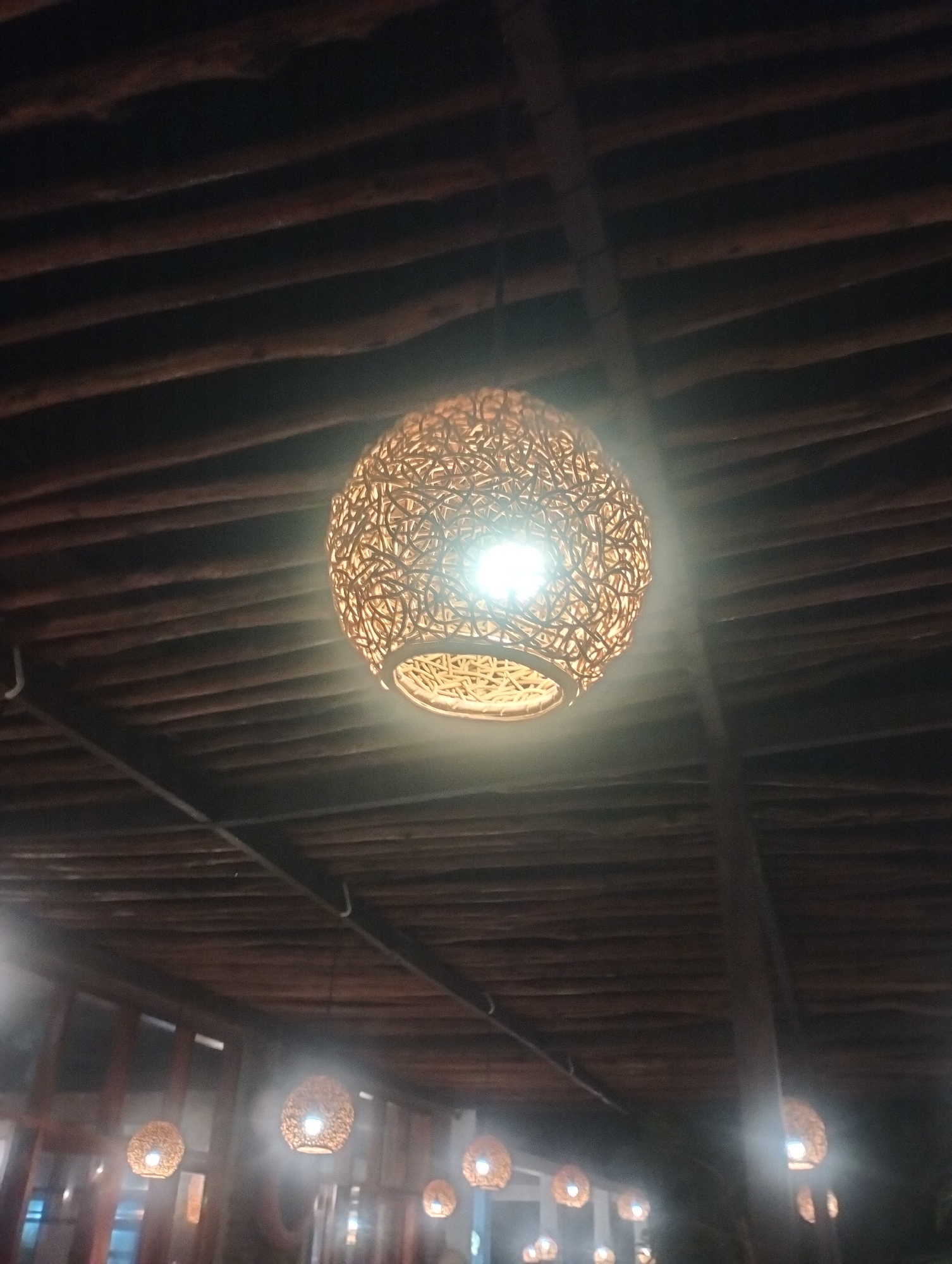 The image displays a spherical light fixture, intricately woven from a natural material like rattan, hanging prominently from a wooden ceiling. The bulb inside the fixture shines brightly, illuminating the woven pattern and casting a warm glow, creating a strong light source in the otherwise dark environment. The ceiling is constructed from numerous parallel, rough-hewn wooden beams or logs, giving the space a rustic and natural aesthetic.
In the background, several other smaller, similarly designed woven light fixtures are visible, softly glowing and contributing to the overall ambiance. These additional lights are arranged in a way that suggests a larger indoor or covered outdoor setting, possibly a restaurant, cafe, or resort common in tropical areas. The dark background confirms that the photograph was taken at night or in a very dimly lit interior.
There are no people or specific activities depicted. No text is visible in the image. The natural materials and warm lighting create a cozy and inviting atmosphere, consistent with the provided location context of Keudekruenggeukueh, Indonesia, which might feature such natural and traditional architectural elements.