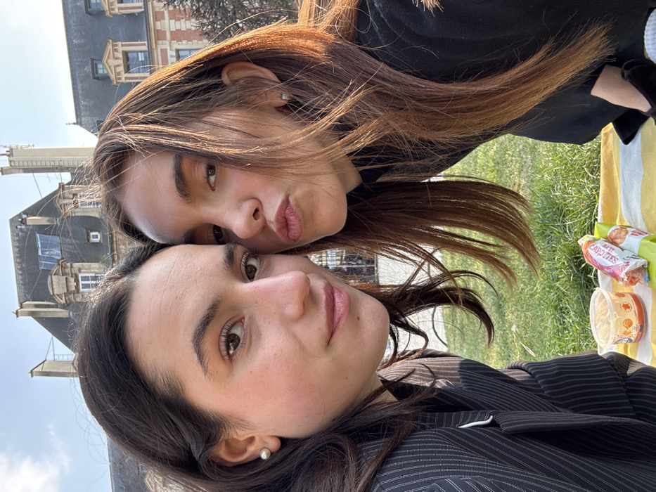 Two young adult females are depicted in a close-up outdoor portrait, likely a selfie, taken in the Place des Vosges, Marais district, Paris, France. The woman on the left, with dark brown hair and a fair complexion, wears a dark pinstriped blazer and a pearl stud earring, presenting a slight smile directly at the camera. The woman on the right, with lighter brown hair and a similar complexion, wears a dark top and a silver hoop earring, making a "duck face" expression. Their heads are angled close to each other, indicating a shared moment.
They are seated on a striped yellow and white blanket placed on green grass. In the immediate foreground, on the blanket, a clear plastic container with a light-colored food item (resembling hummus or a dip) and a red and white packaged snack are visible.
The background reveals classic Parisian architecture under a clear blue sky. Dominant features include light-colored stone facades, some with red brick elements, and characteristic dark grey Mansard roofs with multiple dormer windows, consistent with the historic buildings surrounding the central garden of the Place des Vosges. The scene is illuminated by natural sunlight, casting warm tones.