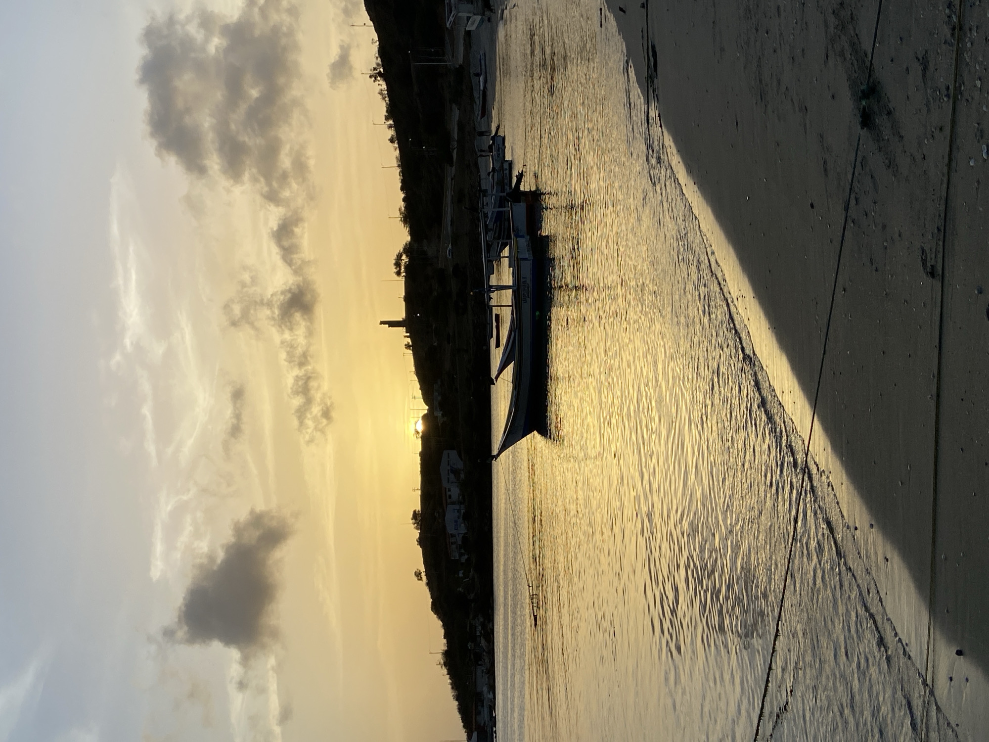 A serene sunset casts a warm, golden glow over the bay in Juan Griego, Venezuela. The sky is painted with hues of yellow and orange, transitioning into a pale blue at the top, with scattered gray clouds adding texture. The sun, partially obscured by a hill, is visible as a bright, radiant disc.
The foreground features a sandy beach with a few scattered shells. A dark shadow stretches across the sand from right to left, likely cast by the rising hill. A dark rope lies on the beach, extending into the water.
The calm water of the bay reflects the warm light of the sunset, creating shimmering patterns on the surface. Several small fishing boats are moored in the water. One boat, closer to the foreground on the right, is partially visible, while another larger boat is anchored further out in the bay. In the mid-ground, more boats are lined up along the shore of the bay.
The background is dominated by a dark, sloping hill covered with sparse vegetation. A few buildings and structures are scattered across the hillside, including what appears to be a lighthouse or tower on the highest point. Power lines and poles are also visible along the crest of the hill, silhouetted against the bright sky. The overall scene is tranquil and picturesque, characteristic of a coastal sunset.
