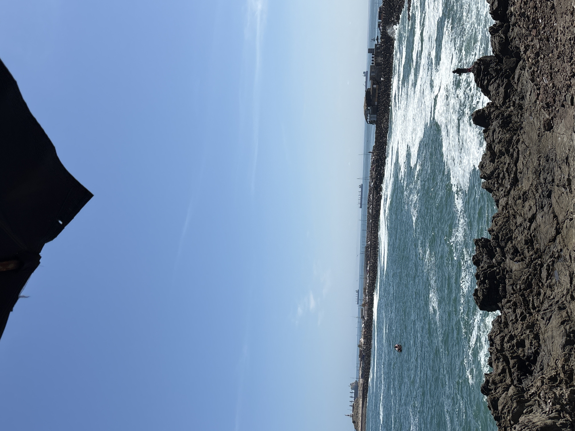 The image displays a vibrant coastal landscape under a bright, clear blue sky. A large expanse of the sky dominates the upper left, partially framed by a dark, irregular object in the top left corner, possibly an awning or roof. The majority of the central and lower-left frame is filled with the dynamic, blue-green ocean, characterized by numerous white-capped waves and foam, indicating active water movement.
On the right side, a rugged, dark-colored rocky coastline or cliff face extends vertically, showcasing uneven, rough textures. A small, dark figure, likely a person, is perched or standing on a prominent rock formation, gazing towards the sea. Further out in the water, closer to the distant horizon, a tiny, dark object, possibly a person on a small watercraft such as a paddleboard or kayak, is visible.
Across the horizon, a long breakwater or jetty stretches horizontally, featuring several structures that resemble buildings or industrial facilities. The overall scene suggests a sunny day with excellent visibility. The strong sunlight and minimal cloud cover point to clear weather. The combination of natural rocky coastline and maritime infrastructure suggests a location like Arica, Chile, which is known for its coastal features. There is no visible text in the image.