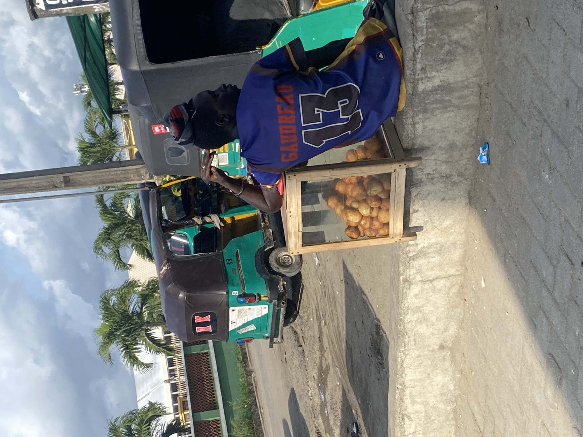 A street scene in Ado, Nigeria. A man in a blue and gold Gaudreau 13 jersey is next to a wooden cart of bread or rolls. The cart is positioned next to a green and brown three-wheeled motorized vehicle (tricycle), possibly a transportation service. Palm trees and buildings are visible in the background under a cloudy sky. The ground is paved, and a small piece of blue trash is visible near the cart.
