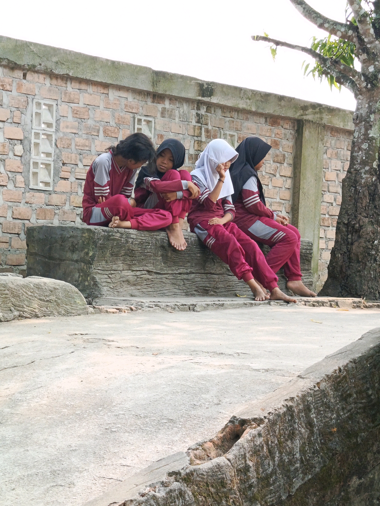 Four young girls in matching maroon and grey tracksuits are sitting on a large, weathered stone slab in front of a brick wall. Three of the girls are wearing headscarves, two black and one white, covering their hair and necks. The girl on the far left is not wearing a headscarf and has her dark hair visible. They appear to be interacting with each other, with two girls looking down, one seemingly talking to another, and the girl on the far right looking slightly away. The stone slab has a rough, textured surface. Behind the girls, a light-colored brick wall with two decorative rectangular openings is visible. To the right of the girls, a portion of a large tree trunk with textured bark is present. The ground in front of the stone slab is a light-colored, possibly concrete, surface with some cracks and debris. The overall environment appears to be outdoors, in a possibly rural or semi-urban setting in Karya Dadi, Indonesia. The sky is overcast and bright.