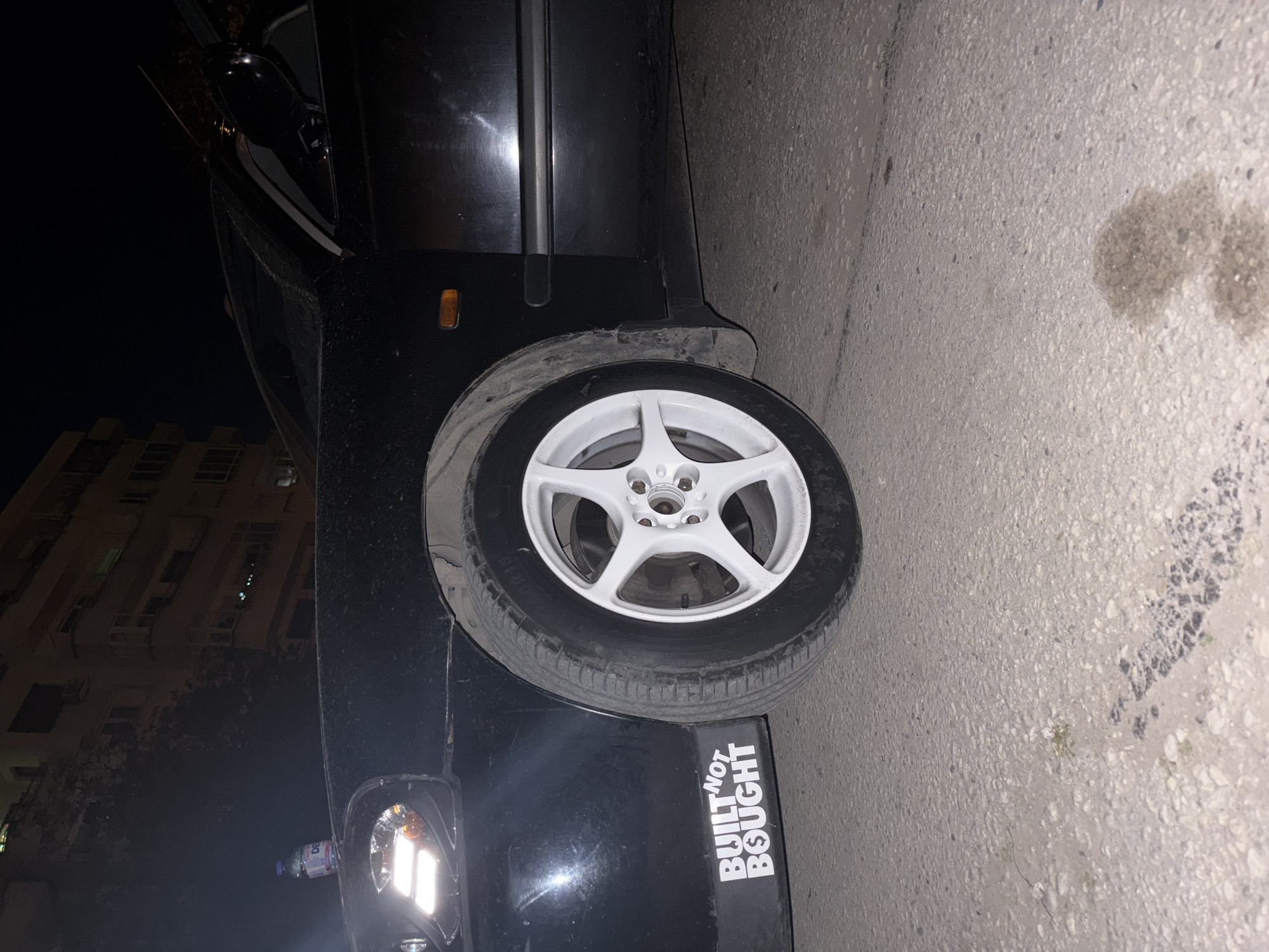 The image shows the rear of a black car parked on a paved street. The car has a white wheel with a black tire. A sticker that reads "BUILT NOT BOUGHT" is on the rear quarter panel. There is a water bottle on the bumper next to the taillight. Across the street, a multi-story building can be seen. The photo may have been taken in Plovdiv, Bulgaria.