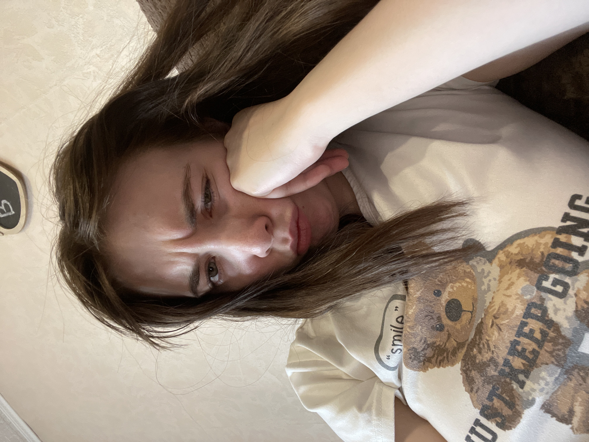 A selfie shows a young woman with long brown hair lying down, her head tilted towards the left. She is wearing a white t-shirt with a teddy bear print and the words "Just keep going" and "smile." Her hand is pressed against her cheek, and her expression is sad, with furrowed brows and downturned lips. The background appears to be a light-colored wall. There is no clear indication in the image to confirm or deny if the location is in Chaikovsky, Russia.