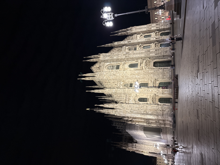 The image depicts the Duomo di Milano, a large Gothic cathedral, illuminated at night in Piazza del Duomo, Milan, Italy. The cathedral's intricate white marble facade, characterized by numerous spires, pinnacles, arched windows, and detailed statuary, is prominently displayed against a dark, clear sky. The left side of the cathedral shows scaffolding or a large white protective covering, suggesting ongoing maintenance or restoration.
The foreground consists of a vast, paved public square composed of light-colored, rectangular stone blocks, some appearing reflective due to illumination. Two tall, dark, ornate lampposts with multiple light globes provide additional illumination to the square; one is positioned on the right and another, smaller one, on the left. A red "M" sign, indicating a Milan Metro subway entrance, is visible near the left lamppost. Low barriers or benches are present in front of the cathedral's base, and a solitary red cone is visible further back in the square.
Approximately 10 to 15 individuals are scattered across the square, appearing as small figures in the distance. They are casually dressed, with some individuals walking, such as two figures on the far left moving away from the cathedral, and others standing. No distinct interactions between people are discernible. Lights from distant buildings are visible on the far right of the scene.