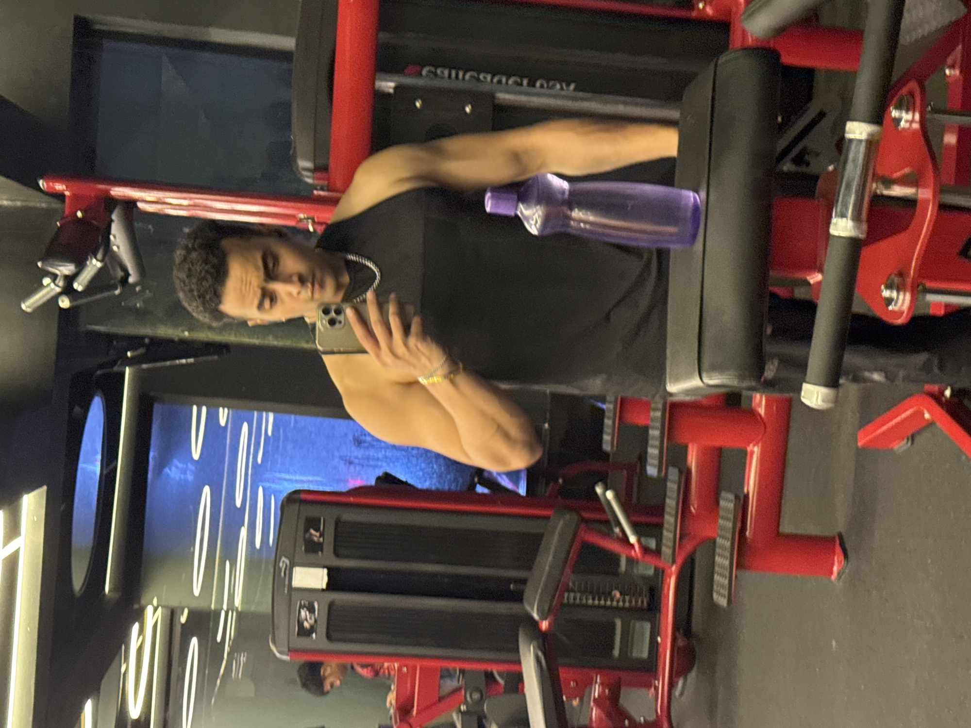 An athletic South Asian man, in his 20s or 30s, with curly dark hair and a goatee, stands in a gym in Kolkata, India, taking a selfie. He is wearing a black sleeveless t-shirt, a silver chain, and a gold bracelet on his left wrist. He holds a silver smartphone in his right hand. To his right, a purple water bottle sits on a black padded seat. The gym equipment is predominantly red and black, with chrome accents. In the background, another person is visible, wearing a red shirt and looking down. The gym appears modern, with bright, linear ceiling lights and a large screen displaying abstract blue imagery. The floor is black rubber matting.