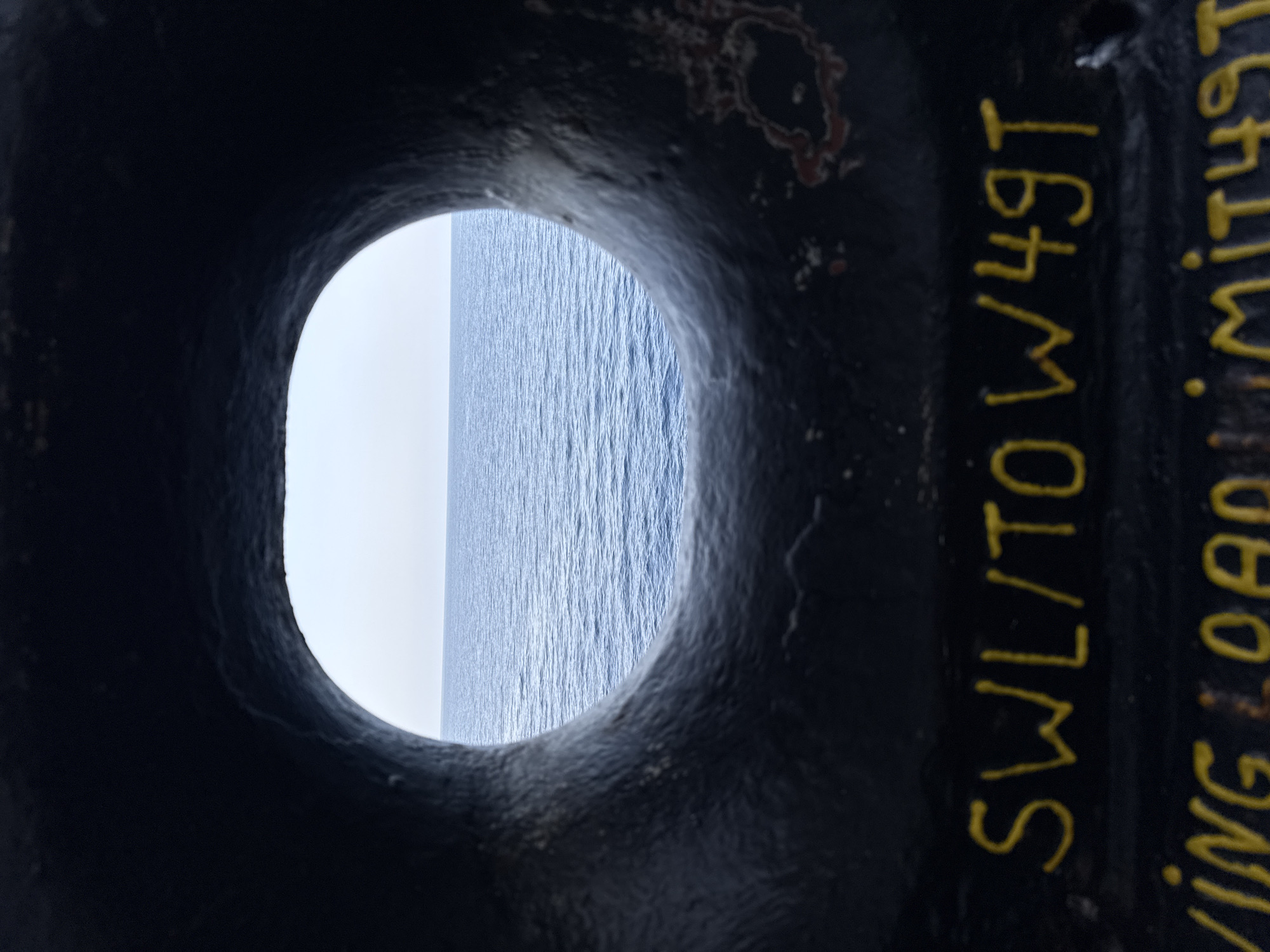 The image is a close-up view through a dark, circular opening, revealing a serene seascape. The opening, likely a porthole or a similar structure on a vessel or offshore platform, frames a vast expanse of blue ocean. The water is calm, with subtle ripples indicating gentle movement, and stretches to a bright, possibly overcast horizon where it meets a pale sky.
On the right side of the frame, the dark, textured surface of the object framing the view is visible. Yellow text is painted onto this surface, oriented vertically and appearing weathered. The legible portions of the text read "SWL/TOW49T" and "VING LOAD LIMIT49". This suggests the object is a piece of industrial equipment, likely related to lifting or towing, with a specified safe working load (SWL) and towing capacity. The presence of the ocean and industrial markings suggests a maritime or offshore setting. The lighting is diffused, indicating it could be daytime with cloud cover, or possibly early morning or late afternoon. There are no people or animals visible in the frame, and the overall mood is one of quiet observation of the natural environment through a man-made aperture. The location context of Águilas, Spain, suggests the scene is set along the Mediterranean coast.