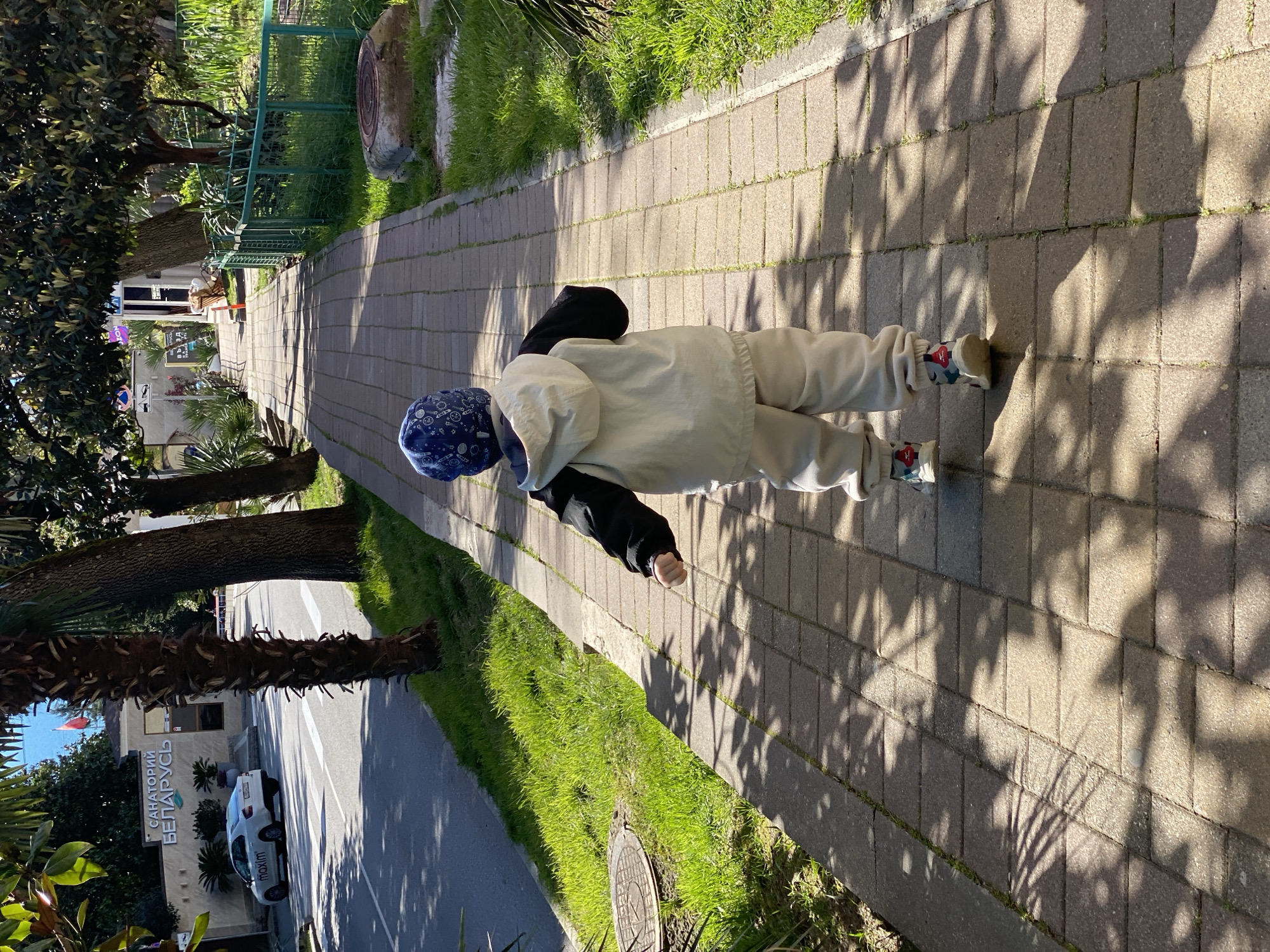 A young child is lying face down on a brick sidewalk next to a grassy area in what appears to be a city park or promenade. The child is wearing a light-colored jacket, pants, and sneakers with a blue patterned hat. Shadows from trees are cast across the sidewalk. A street with a white car parked next to a building is visible in the background. A sign written in Russian on the building reads "SANATORIY BELARUS". This indicates the location is near the Sanatorium Belarus in Sochi, Russia.