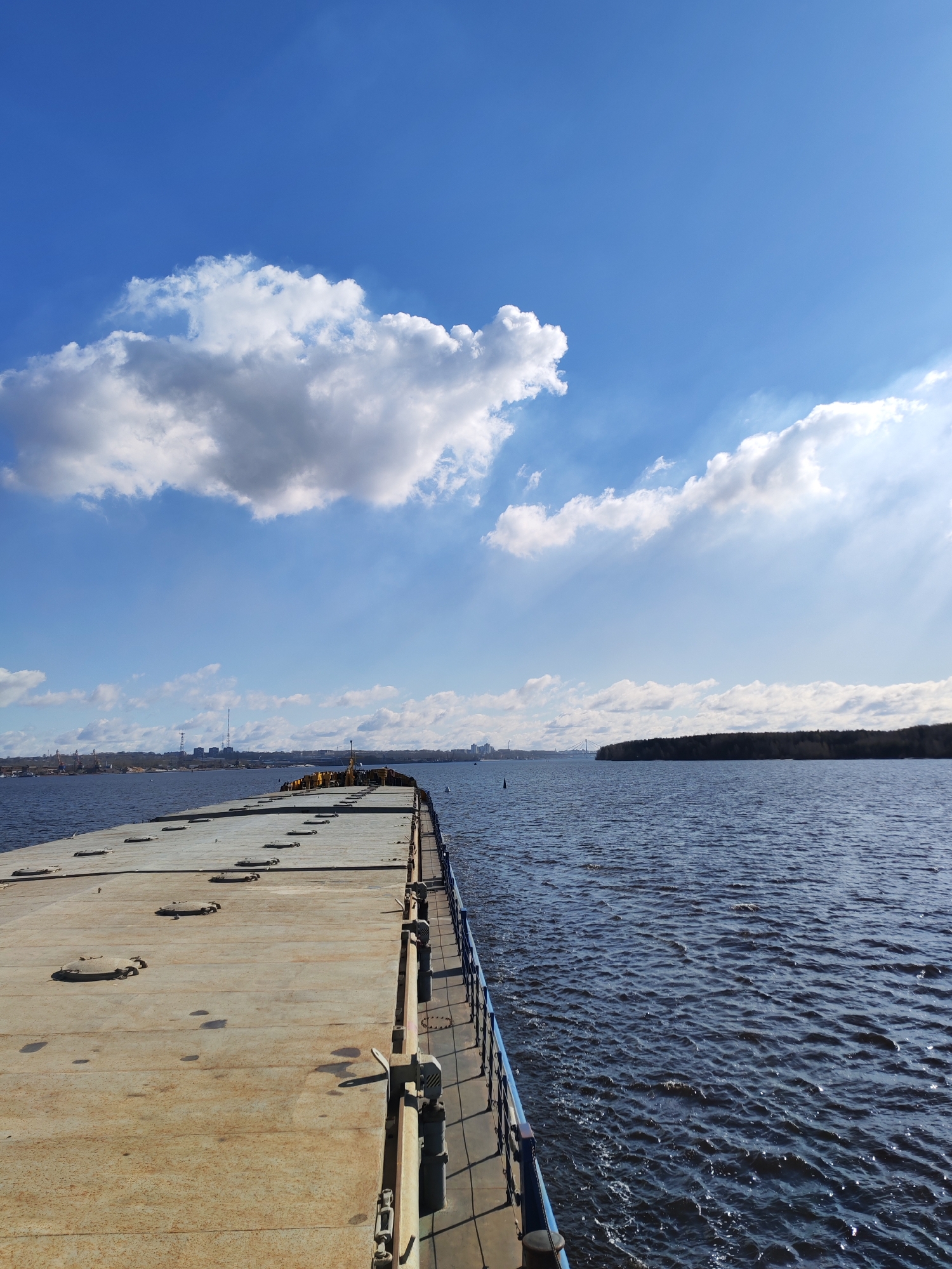The image depicts a barge on the Sheksna River in Cherepovets, Russia. The barge is flat and grey, with round access hatches on its surface. A blue railing runs along the edge of the barge. The river is dark blue with small waves. In the distance, on the left bank, are industrial structures and buildings. The right bank is covered with dense green trees. The sky is blue with fluffy white clouds. Sunlight streams down from the upper right corner of the frame.