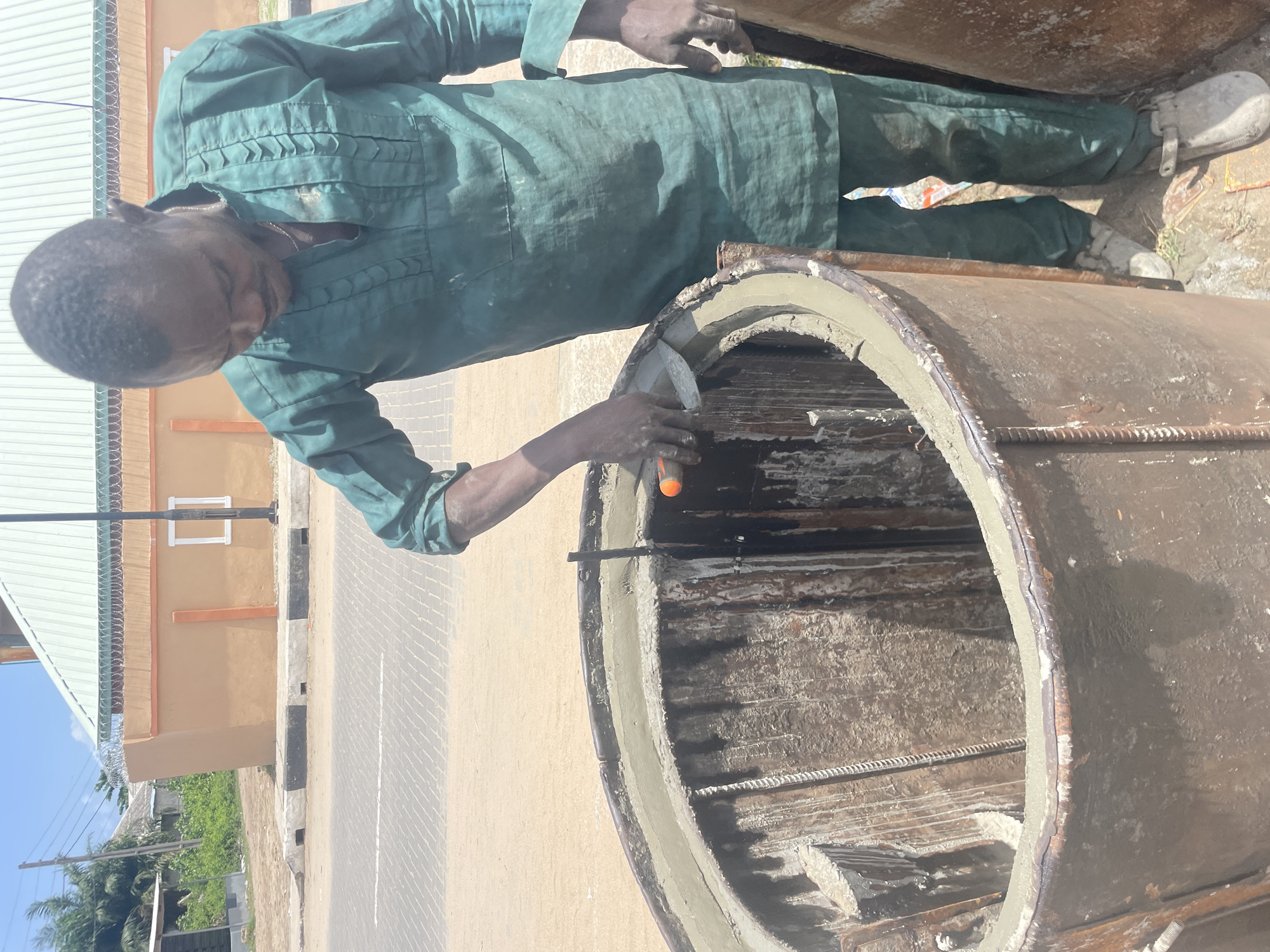 The media file displays an outdoor scene in Badagry, Nigeria, featuring a man engaged in construction or craftsmanship. The primary subject is an adult man, likely a worker or artisan, dressed in a dark green coverall. He is focused on applying and smoothing a layer of wet mortar or cement along the top rim of a large, robust cylindrical mold, possibly for a concrete pipe, well ring, or similar precast concrete structure. He holds a trowel or spatula in his right hand, precisely shaping the material. The inside of the mold shows rough wooden or metal surfaces and rebar reinforcement, indicating it's a formwork for casting concrete.
The setting appears to be a sunny day, with bright, direct light creating distinct shadows, suggesting it's daytime. In the background, there's a light brown or orange-colored building with a white window frame and a light green corrugated metal roof. A fence topped with barbed wire runs along the side of this building. Beyond the structure, a paved road or pathway is visible, marked with white and black curb stones. Lush green foliage, including palm trees, is present further in the distance under a clear blue sky. The overall impression is one of a busy, practical working environment under pleasant weather conditions. No visible text is present in the image.