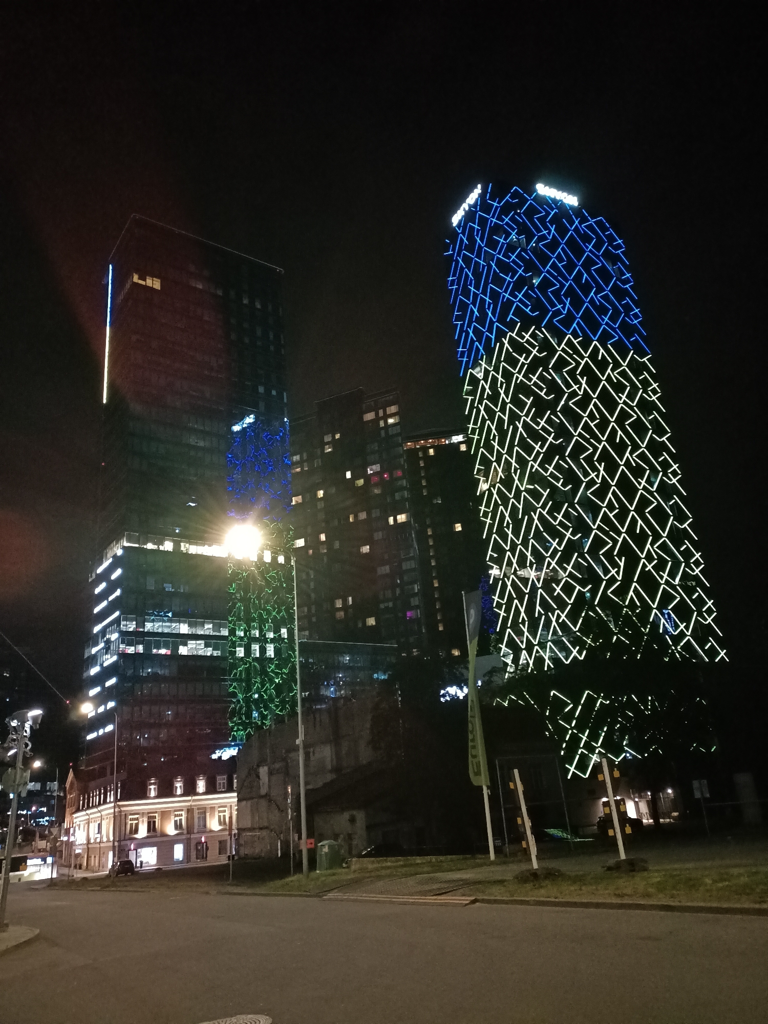 The image captures a night scene in Tallinn, Estonia, specifically focusing on the modern architecture of the T1 Mall of Tallinn. The foreground shows a paved street and a grassy area, with streetlights and a traffic signal visible on the left. Dominating the background are two distinct skyscrapers. The left skyscraper is a tall, dark building with illuminated windows and vertical strips of blue and green lights. The right skyscraper is a more uniquely designed building, featuring a facade covered in a geometric pattern of bright blue and green lights that illuminate the night sky. In between the skyscrapers, a lower, older building with illuminated windows is visible, alongside some darker, industrial-looking structures. The overall impression is of a modern urban landscape at night, illuminated by the vibrant lights of the buildings and street infrastructure.