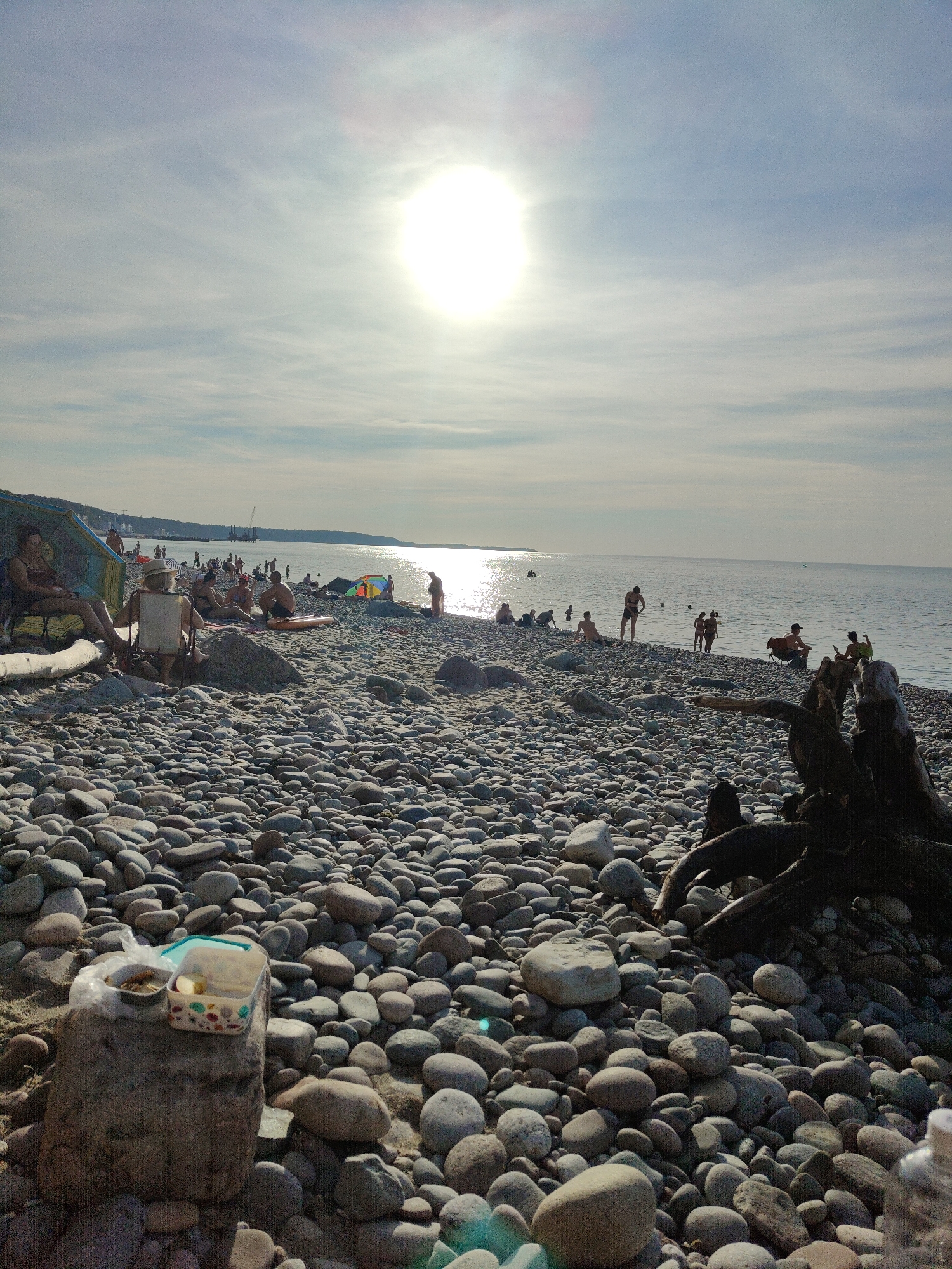 The image depicts a lively beach scene in Pionersky, Russia, under a bright, hazy sun. The foreground is dominated by a vast expanse of smooth, grey pebbles. A large rock in the lower left corner supports an open lunchbox with visible food items and a plastic bag. Further back, a group of people are scattered across the pebble beach, some seated under a blue beach umbrella, others relaxing on towels or chairs. Several colorful beach tents are visible.
Beyond the immediate beach area, the sea stretches out, its surface shimmering with reflected sunlight. People are dotted along the shoreline and in the shallow water, engaging in various beach activities. In the distance, the coastline is lined with trees and structures, and a construction crane is visible against the horizon, suggesting ongoing development. The overall atmosphere is one of a pleasant summer day at the beach, with people enjoying the sun and the sea.