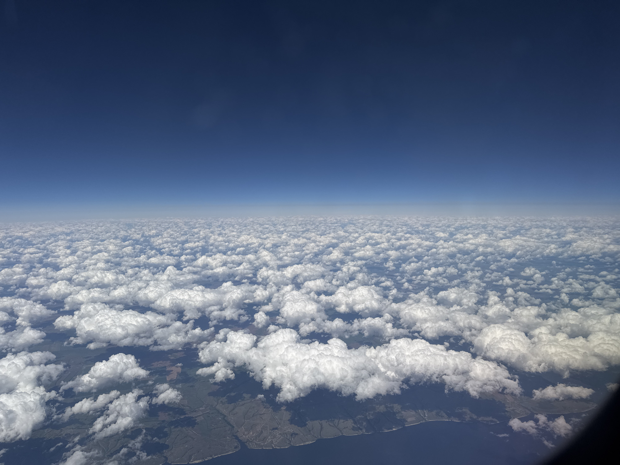 The image depicts a view from above a layer of cumulus clouds. Above the clouds is a clear, deep blue sky. Below the clouds, land is visible, punctuated by bodies of water. Based on the prompt including the city of Izhevsk, Russia, it is probable that the land is in the vicinity of Izhevsk and the water bodies are the Votkinsk Reservoir on the Kama River, however, without GPS coordinates it is not possible to confirm this hypothesis with certainty. No people or subjects are visible. The camera is inside an airplane.
