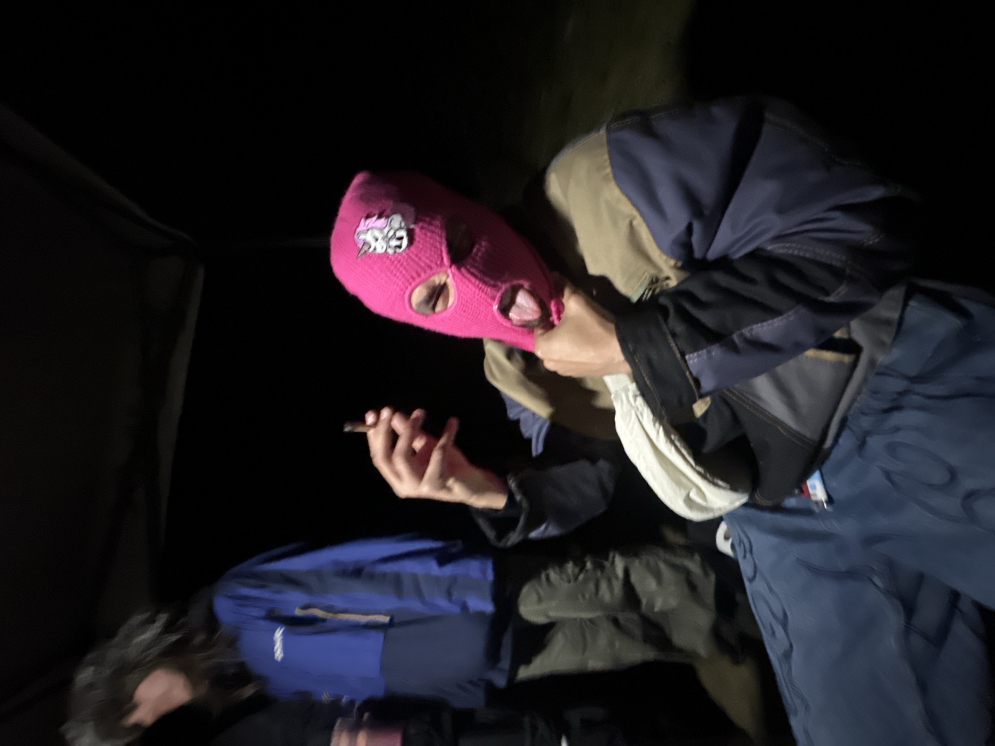 The image shows two individuals outdoors at night. One person wears a pink balaclava with a logo and holds a lit cigarette in their right hand. They wear a multi-toned jacket with blue, black, beige, and white panels, and blue pants. The other person lies down, partially visible and wearing a blue jacket. The background is dark, obscuring details but suggesting a wooded or natural environment. The presence of a tent suggests the location is a campsite. Specific location details within Auvers-Saint-Georges, France, are not discernible from the image.