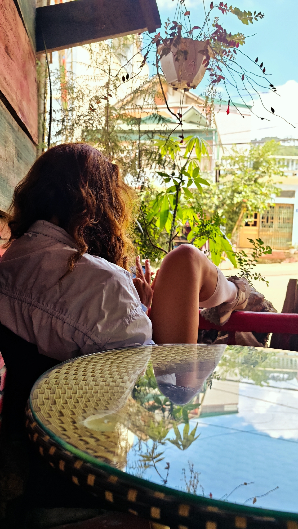 A young person with reddish-brown curly hair sits at a round table in an outdoor setting, possibly a café. The individual wears a light-colored jacket. One leg is visible, propped up on a red railing. A hanging plant is seen above, and foliage and building facades are visible in the background. The table is made of wicker with a glass top, reflecting the sky and the surrounding environment. The scene appears to be set in Đồng Văn Commune, Vietnam.
