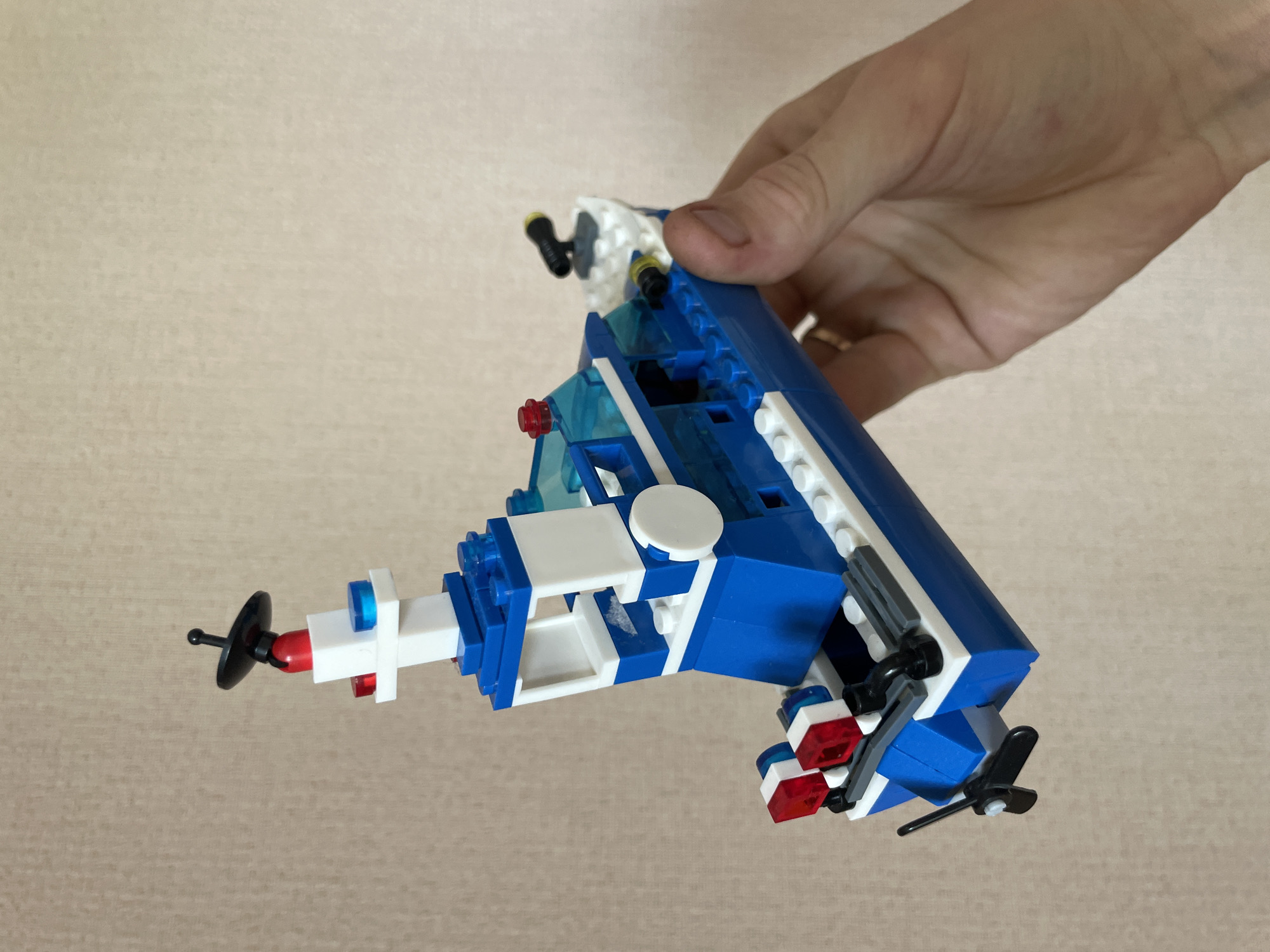 A person's hand is holding a blue and white Lego boat with a propeller and red and blue lights. The boat has a black antenna and two black cannons on the back. The background is a textured beige wall. There is no indication of the city of Laishevo, Russia in the image.