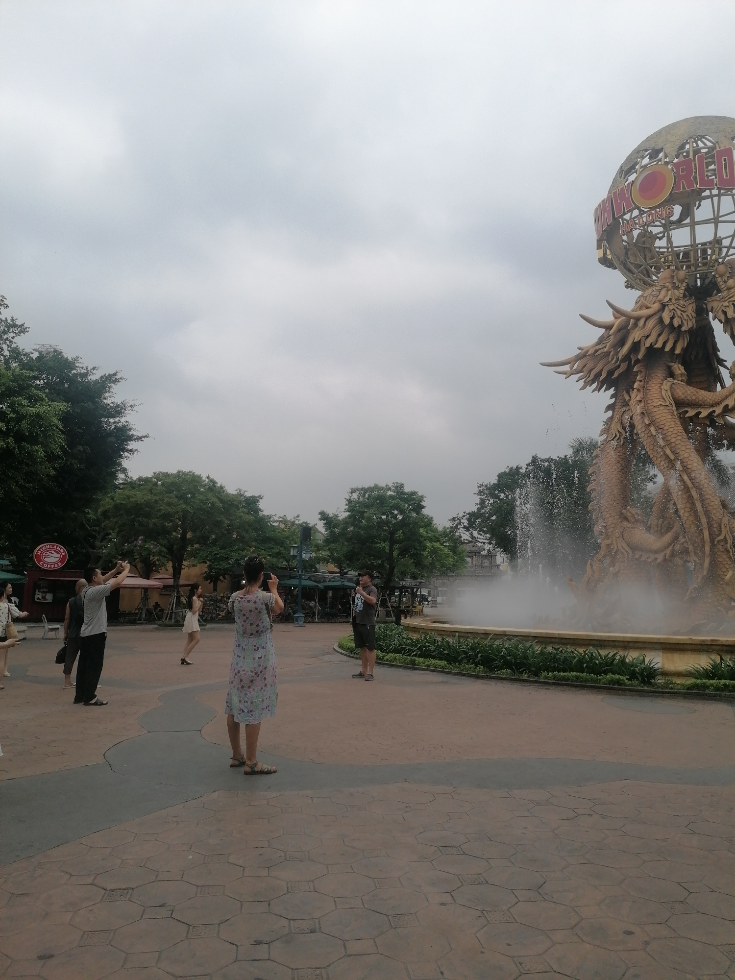 The image captures an outdoor scene at what appears to be an amusement park or tourist attraction, specifically Sun World Ha Long in Bai Chay Ward, Vietnam. The sky is overcast and cloudy, suggesting a dull but bright daytime.
In the foreground and mid-ground, several people are visible. Most are standing, facing towards a large, prominent golden dragon statue integrated with a fountain on the right side of the frame. Many of them are holding up smartphones, seemingly taking pictures or videos of the impressive monument.
The central feature of the scene is a colossal, ornate golden dragon statue, coiling around itself and serving as the base for a large, open-work globe structure. Water sprays vigorously from the fountain surrounding the dragon, creating a misty effect at its base. On the globe, the text "SUNWORLD HALONG" is clearly visible, identifying the location.
To the left of the frame, lush green trees line the background. Another notable object is a red circular sign with "HIGHLANDS COFFEE" written on it, indicating a nearby cafe. The ground is paved with reddish and gray interlocking geometric patterns. The atmosphere appears to be that of tourists exploring and documenting a significant landmark.