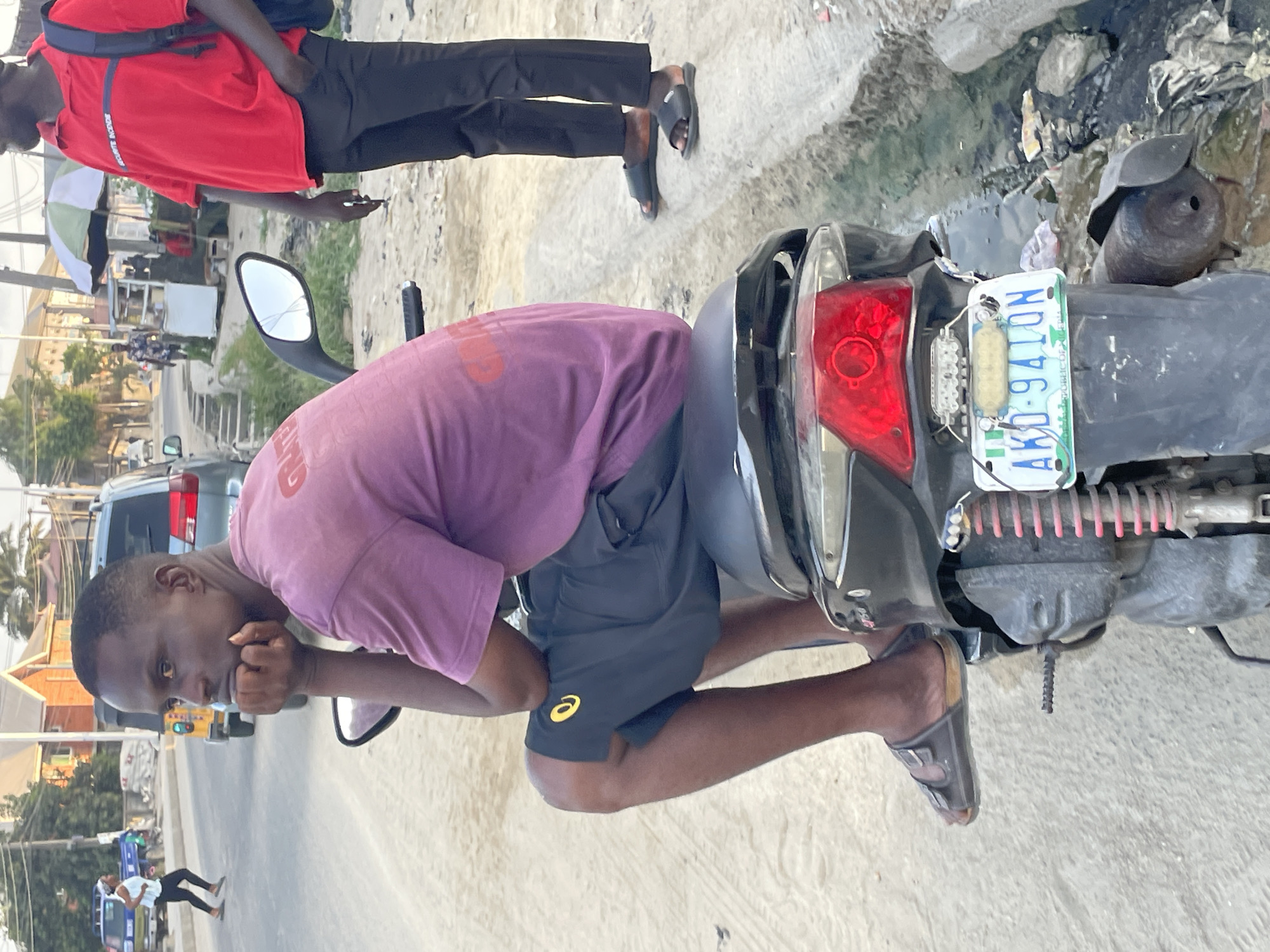 The image depicts two men in what appears to be the city of Ado, Nigeria. One man wears a red shirt and black pants and is standing. Another man in a mauve shirt and black shorts sits on a black motorcycle, possibly examining it. The motorcycle has a license plate that reads "AKO-941QN". The surrounding environment shows a street scene with buildings, pedestrians, and parked vehicles.