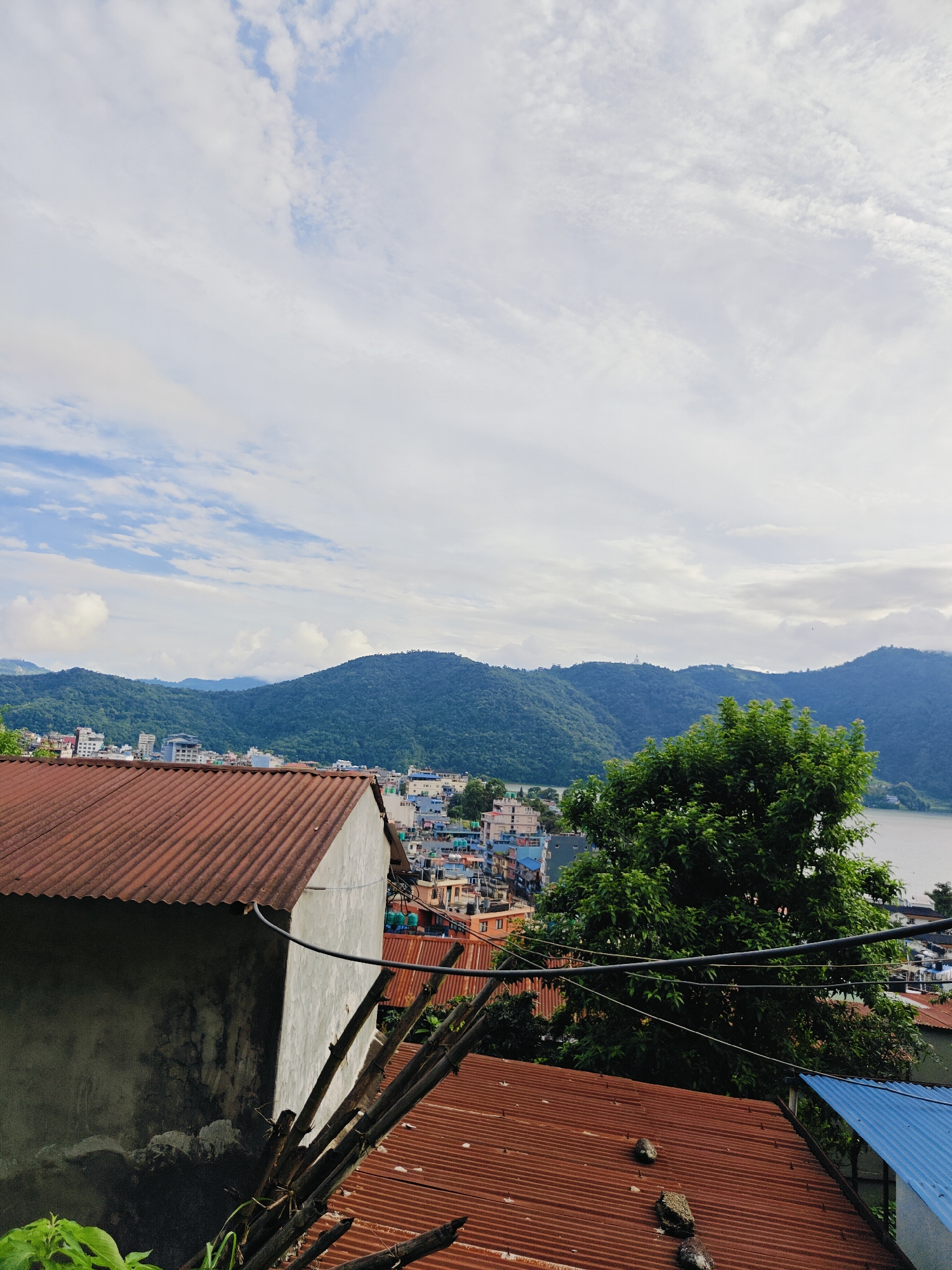 The image depicts a panoramic view of Pokhara, Nepal, from an elevated vantage point. In the foreground, two rustic, corrugated metal roofs are visible, one a reddish-brown hue and the other a more weathered, darker shade, with some stones scattered on the surface of the latter. A partially visible white concrete structure, likely a building, abuts the reddish roof, showing signs of age and weathering. Several bamboo poles are propped against this structure, adding to the scene's rustic charm.
Beyond the immediate foreground, a densely populated cityscape unfolds. Buildings of various colors, primarily blues, whites, and oranges, are clustered together, forming a vibrant urban tapestry. The city appears to be built on a hillside, with structures cascading down towards a body of water. In the mid-ground and background, lush, green, tree-covered hills rise majestically, framing the city and creating a striking contrast between the urban development and the natural landscape. A large, verdant tree stands prominently in the mid-right of the image, its dense foliage contributing to the overall sense of natural beauty.
In the distance, a serene lake or reservoir is visible, its calm surface reflecting the overcast sky. The sky itself is a canvas of white, wispy clouds against a pale blue backdrop, suggesting a pleasant, possibly humid, day. The overall atmosphere of the image is one of a bustling city nestled harmoniously within a breathtaking natural environment.