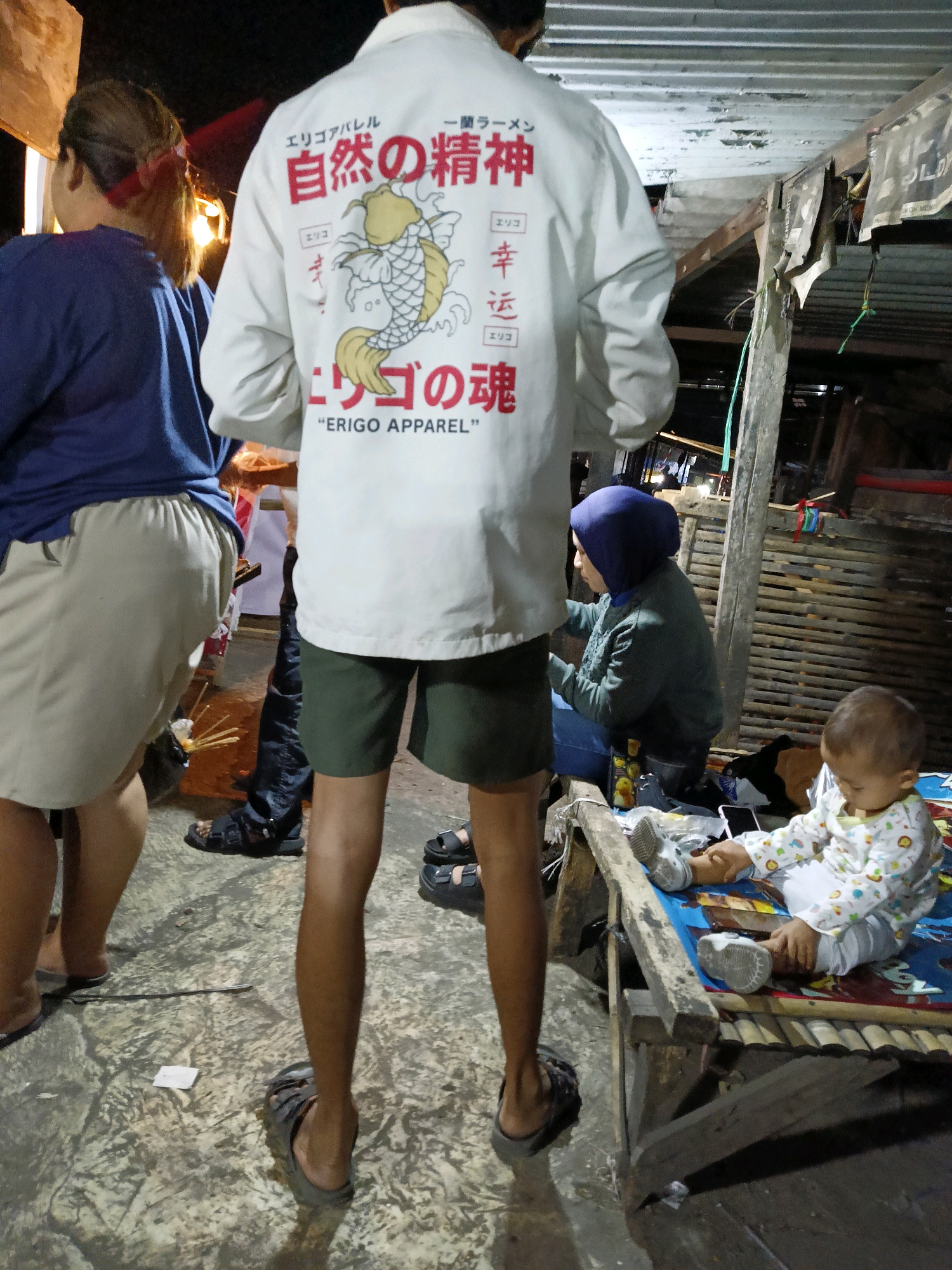 The image shows a group of people gathered outdoors at night, possibly at a market or street food stall in Dompol, Indonesia. In the foreground, a person wearing a white jacket with Japanese characters and a koi fish graphic on the back stands facing away from the camera. They are wearing green shorts and dark sandals. To the left, another person in a dark blue shirt and a light-colored skirt is partially visible. Further back, a woman wearing a blue headscarf and a green jacket is seated, and a young child in a light-colored outfit is sitting on a wooden structure. The environment appears to be a rustic, open-air setting with wooden structures and corrugated metal roofing visible in the background. The ground is a textured concrete surface. The lighting suggests it is nighttime, with artificial lights illuminating the scene.