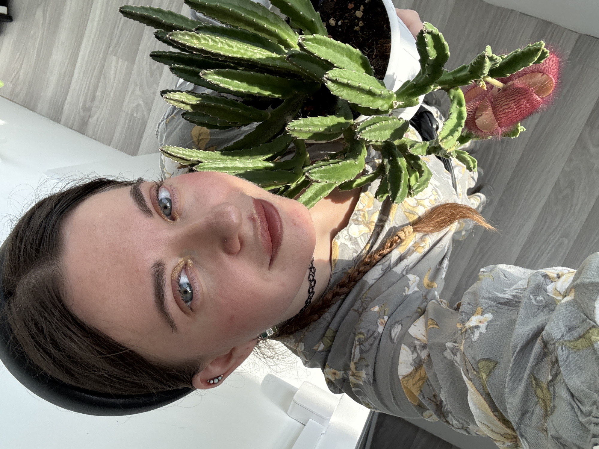 A high-angle, medium shot captures a young Caucasian woman holding a potted cactus. The woman is positioned slightly left of center, looking directly at the camera with a gentle smile. She has light skin, blue-green eyes, and dark brown hair pulled back with a wide black headband. Her makeup includes shimmering gold eyeshadow and subtle lip color. She wears a grey dress with a floral pattern featuring yellow and white flowers. A black choker necklace is visible, and her hair is styled in a braid that extends over her shoulder. She has multiple piercings in her left ear.
The cactus, held in a white pot, is to the right of the woman and fills a significant portion of the frame. It has thick, green, ribbed stems with small white spines. A large, pinkish-red flower with a fuzzy texture and intricate markings is attached to one of the stems, hanging downwards.
The background consists of a white wall on the left and a grey wooden floor with horizontal planks on the right. Sunlight casts shadows on the floor, indicating a well-lit environment. The scene appears to be indoors, possibly in a home. No specific city or location details are discernible from the image itself.