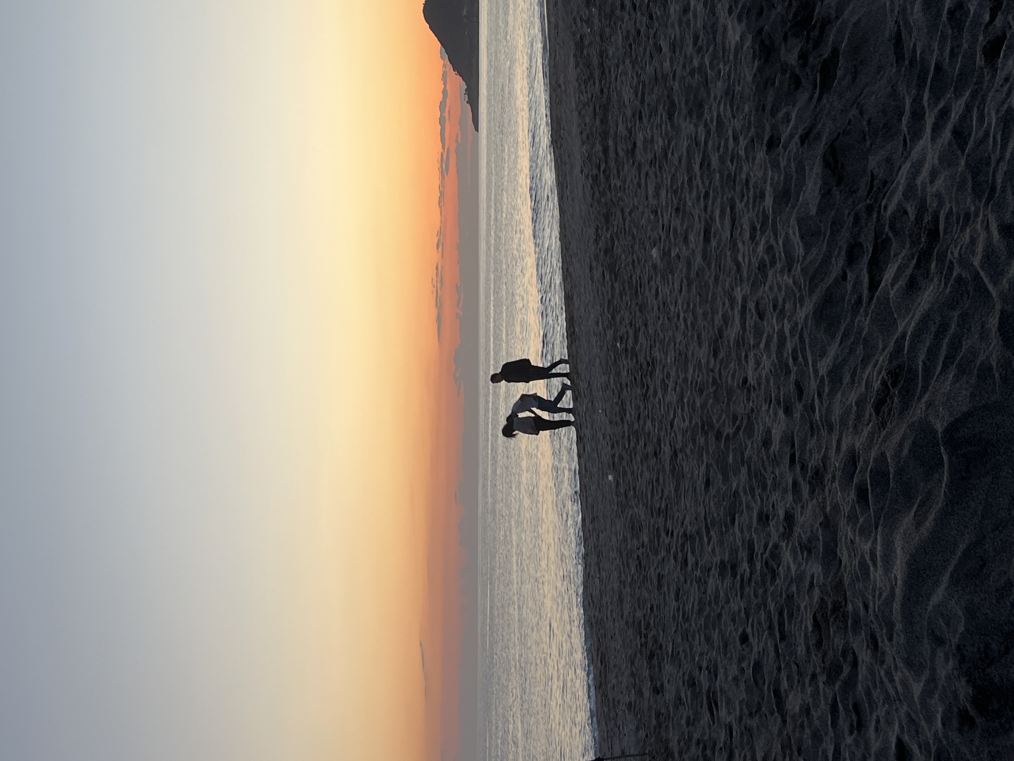 The image captures a coastal scene at dusk, likely in Alanya, Turkey, given the sandy beach and potential mountain silhouette in the background. The photograph is oriented vertically, with the sky transitioning from light blue to orange in the upper left. Silhouettes of two people stand near the water's edge; one appears to be taller than the other, possibly an adult and a child. They may be walking or interacting, but the details are obscured. The right side of the image is dominated by dark sand with distinct textures. The sea occupies the middle ground.