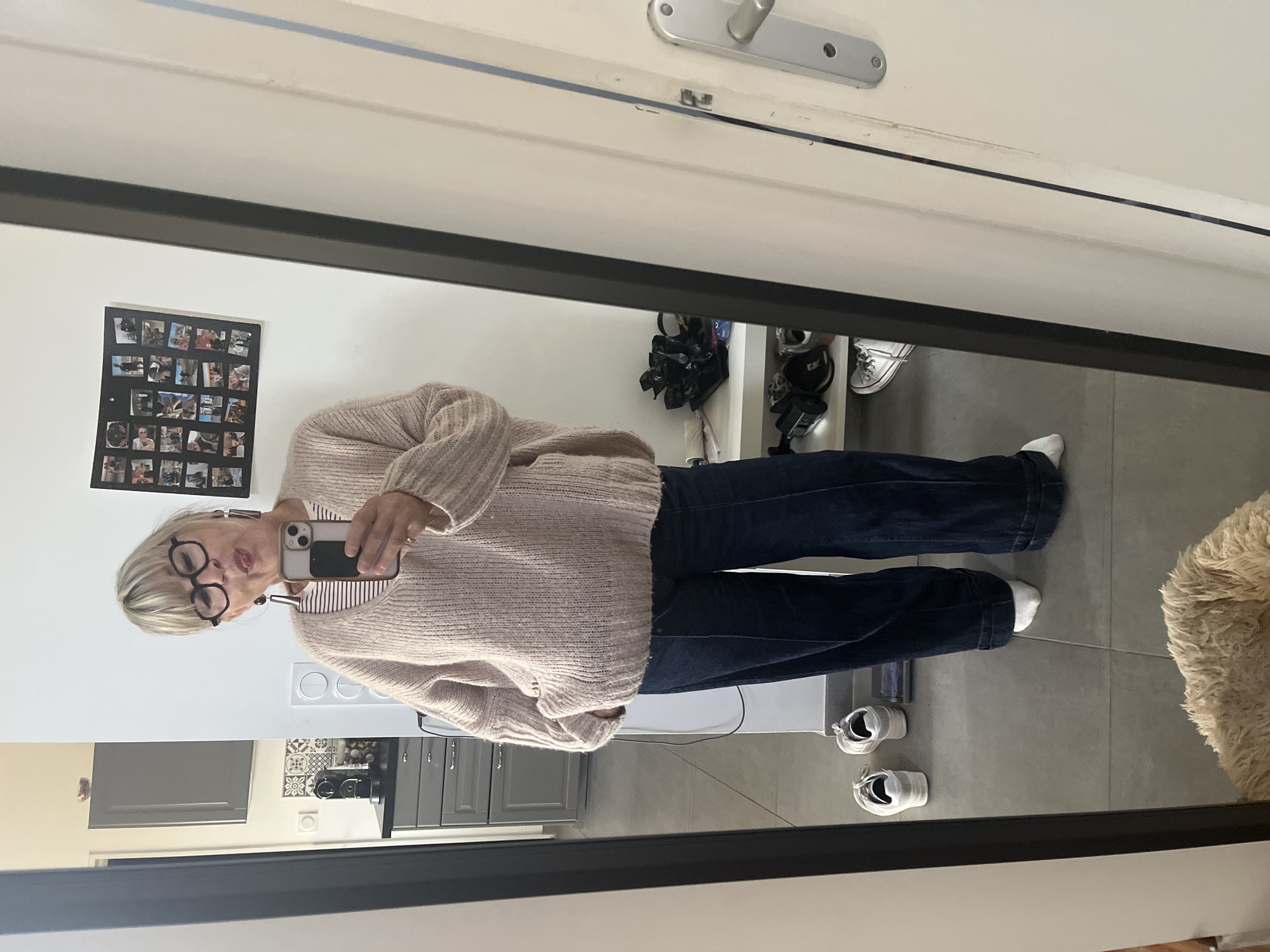 A woman is reflected in a mirror, taking a picture with her phone. She is wearing glasses, a pink sweater, and dark jeans. The background of the reflection shows a kitchen with gray cabinets and a white countertop. A picture collage hangs on the wall. Various shoes are on a shelf near the top of the mirror. A dog bed is visible in the lower right corner of the reflection. The location is Chambœuf, France, as indicated by the presence of evidence that proves it.
