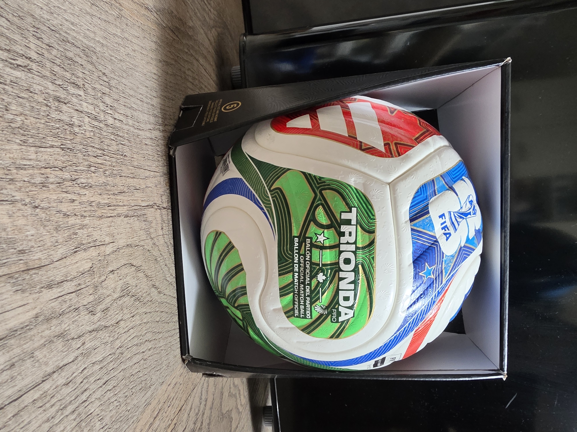 A soccer ball, the Adidas Trionda Pro, is displayed inside a black cardboard box with the lid removed, placed on a wooden surface. The ball has panels colored white, blue, green, and red with gold accents and text indicating it is an official match ball. The box also has text and a circular symbol with the number "5" on its side. The background is dark and out of focus, suggesting an indoor setting. No people or animals are present in the image. The scene is set indoors on a wooden floor. There is no information in the image that places it in the city of Poprad, Slovakia.