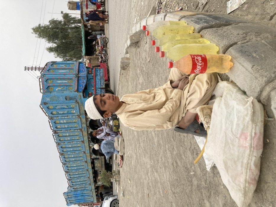 The image depicts a roadside scene in Taftan, Pakistan, characterized by commercial activity and transportation infrastructure under an overcast sky.
In the foreground, a young male, appearing to be a pre-teen or early teen, sits cross-legged on a dusty ground surface. He wears a light tan shalwar kameez and a white knitted prayer cap (topi). His expression is neutral as he looks slightly to his right. His hands are clasped in his lap, and dark sandals are positioned near his left foot. To his right, six transparent plastic bottles containing a yellow liquid, consistent with fuel, are arranged on a grey concrete curb. An orange-tinted plastic bottle, labeled "Coca-Cola" but also appearing to contain a yellow liquid, stands in front of the others. To the boy's left, a white woven bag lies on the ground, with a yellow utensil partially visible.
In the midground and background, a large, elaborately decorated blue cargo truck dominates the left side. The truck features intricate Pakistani truck art, including vibrant patterns and script. Two adult males are seated on the ground near the left side of this truck, appearing to be resting or conducting a transaction. One wears a white cap, the other a reddish scarf. Further to the right, beyond the truck, several motorcycles are parked near a makeshift market stall or shop, which has a green tarp awning. Multiple individuals, dressed in traditional attire, are visible standing and interacting near this stall. Tall, slender trees and utility poles with power lines are present in the far background. The overall environment is dusty, suggestive of a busy transit or market area.